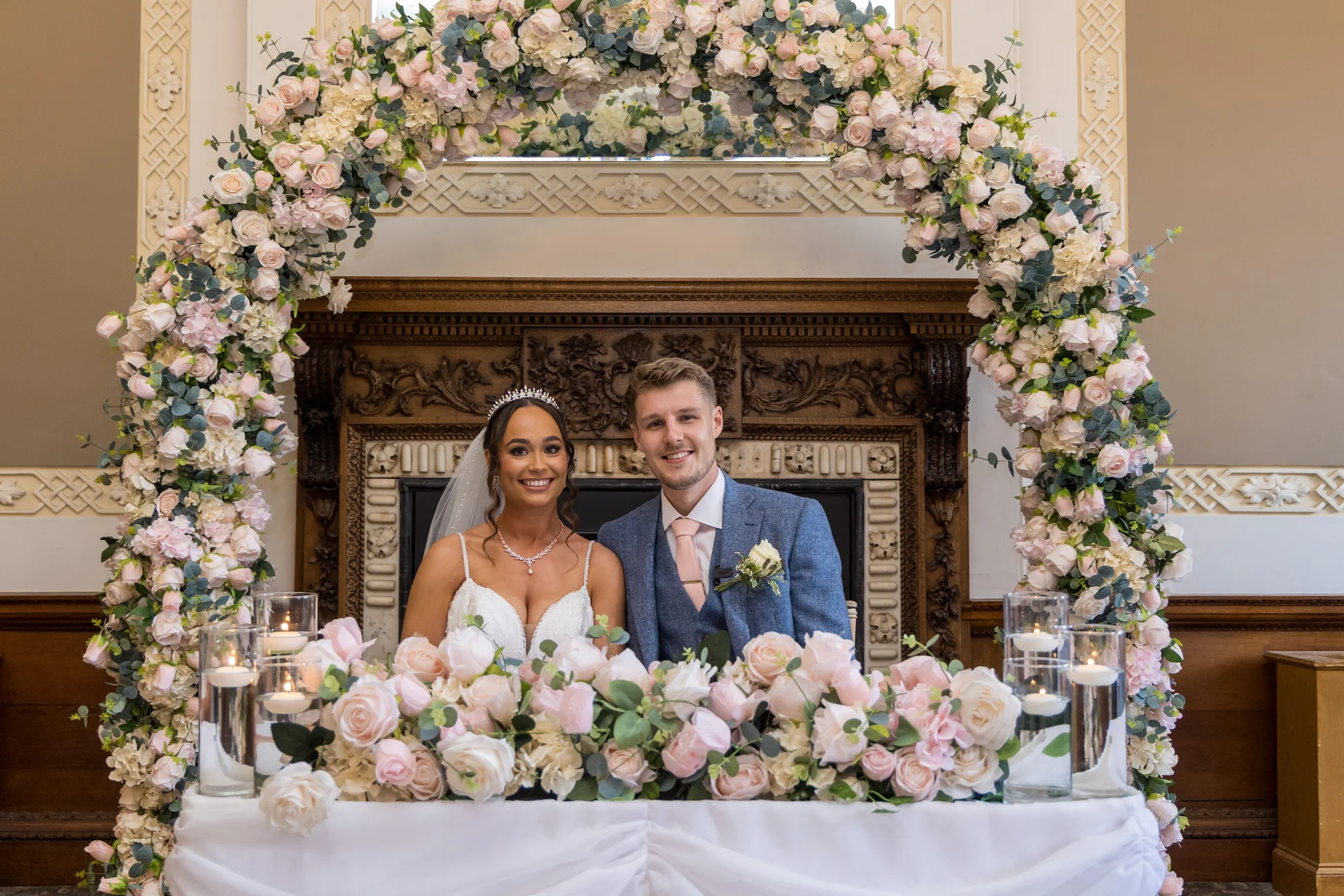 A smiling bride and groom sit together at a sweetheart table framed by a large floral arch and an ornate wooden fireplace.