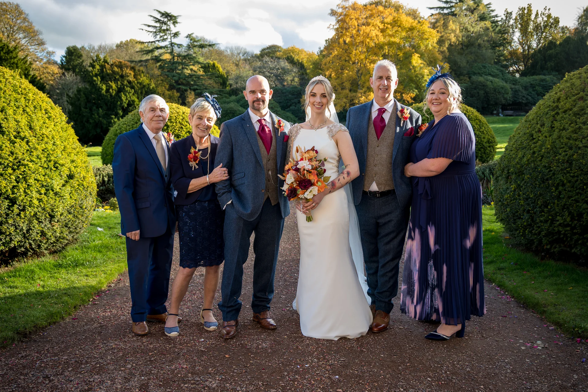A newlywed couple and their family members pose together on a pathway lined with manicured hedges during an autumn wedding.