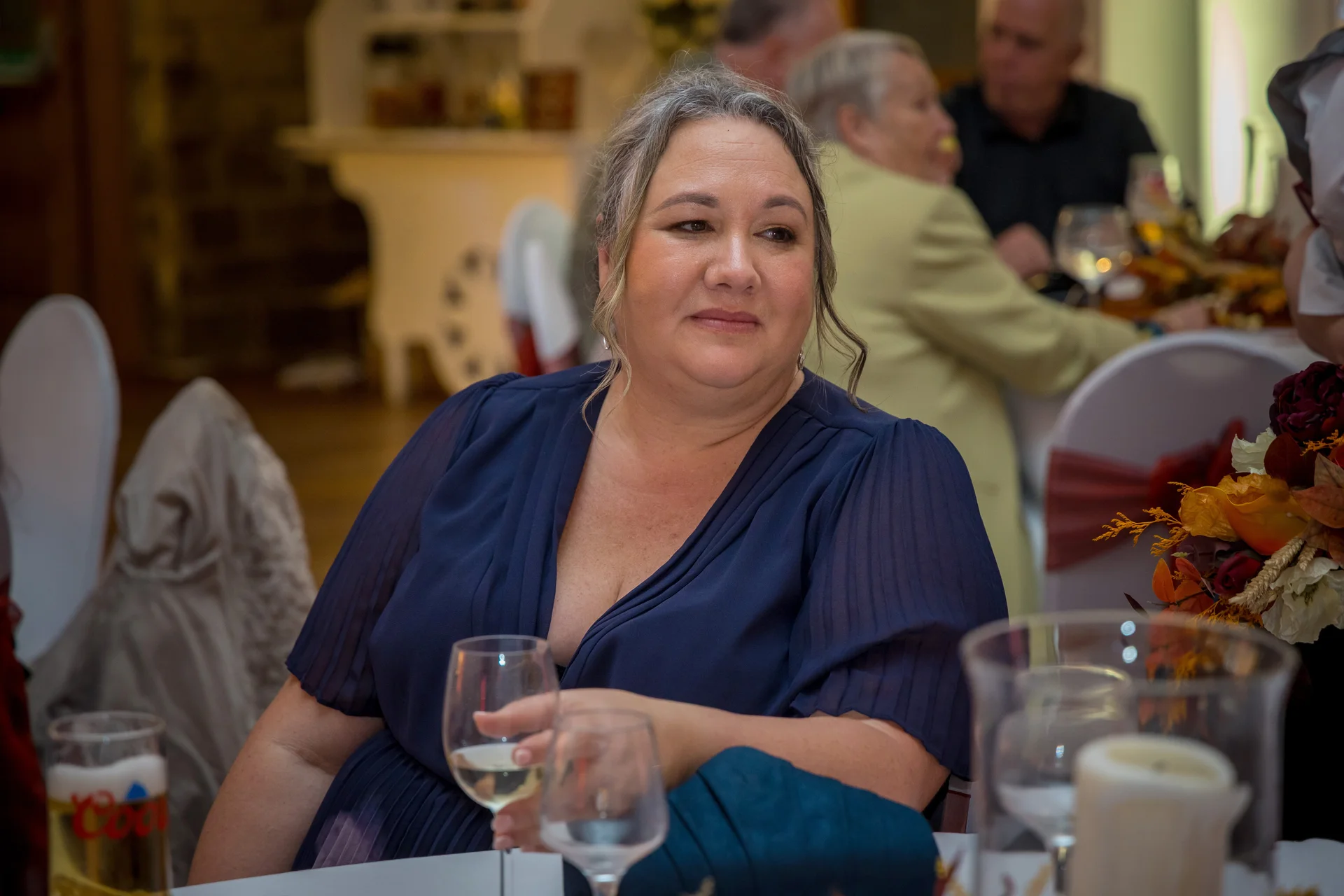 A wedding guest in a dark blue pleated dress sits at a reception table, holding a wine glass and looking attentively off-camera.