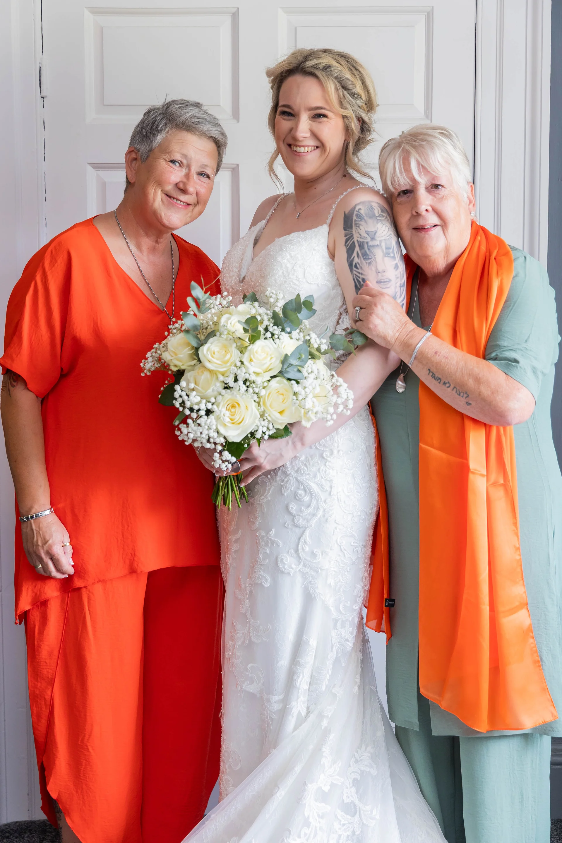 A smiling bride in a lace gown holding a white rose bouquet stands proudly between two family members indoors.