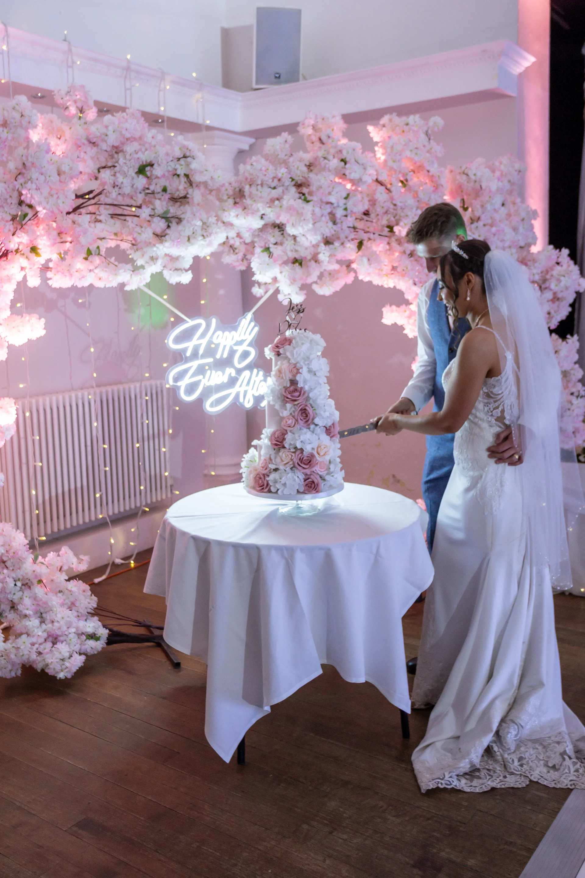 A newlywed couple cuts a tiered floral wedding cake together beneath a glowing neon sign and an elaborate arch of pink blossom.