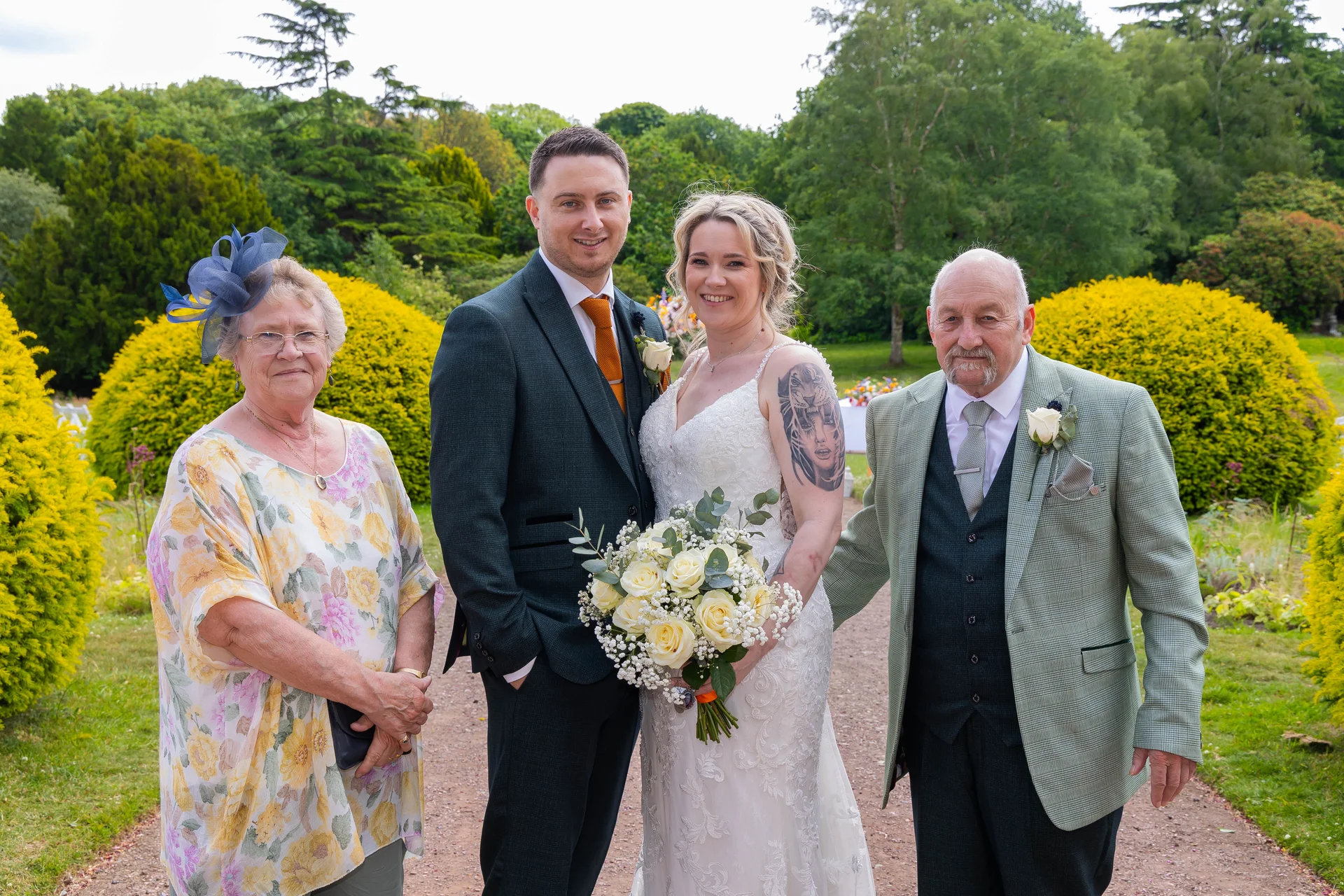 The bride and groom pose outdoors in a lush garden setting alongside family members, holding a white rose bouquet.