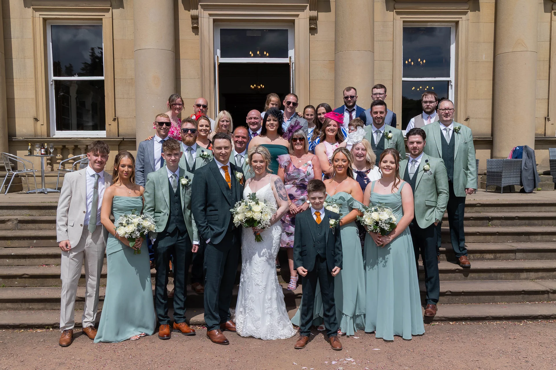 The wedding party and guests pose for a large formal group photograph on the stone steps outside a grand country house venue.