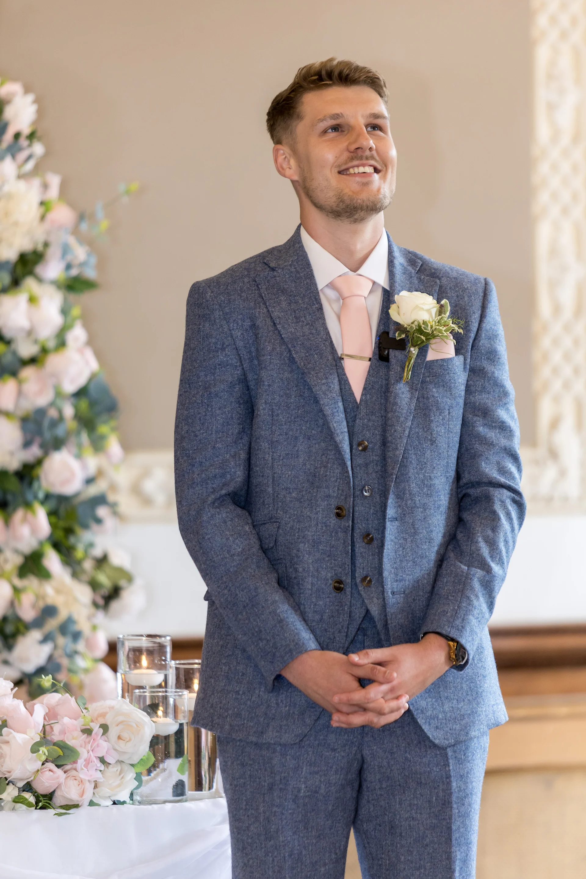 A smiling groom wearing a blue tweed three-piece suit with a pink tie and white rose buttonhole stands at the altar.
