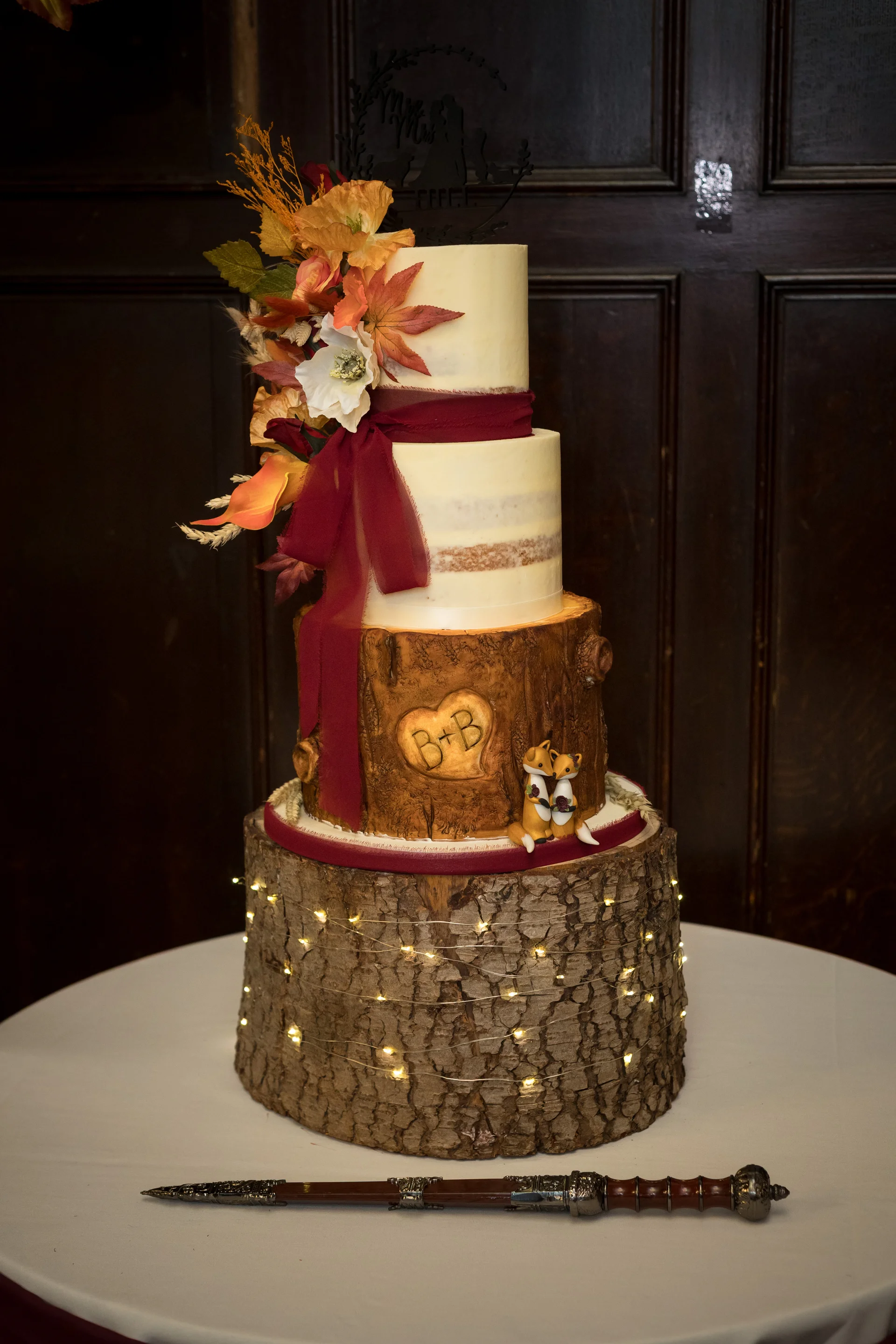 A stunning multi-tiered wedding cake featuring semi-naked tiers, a faux-wood tier with carved initials, autumnal foliage, a burgundy ribbon, and cute fox figurines, displayed on a tree stump base wrapped in fairy lights.