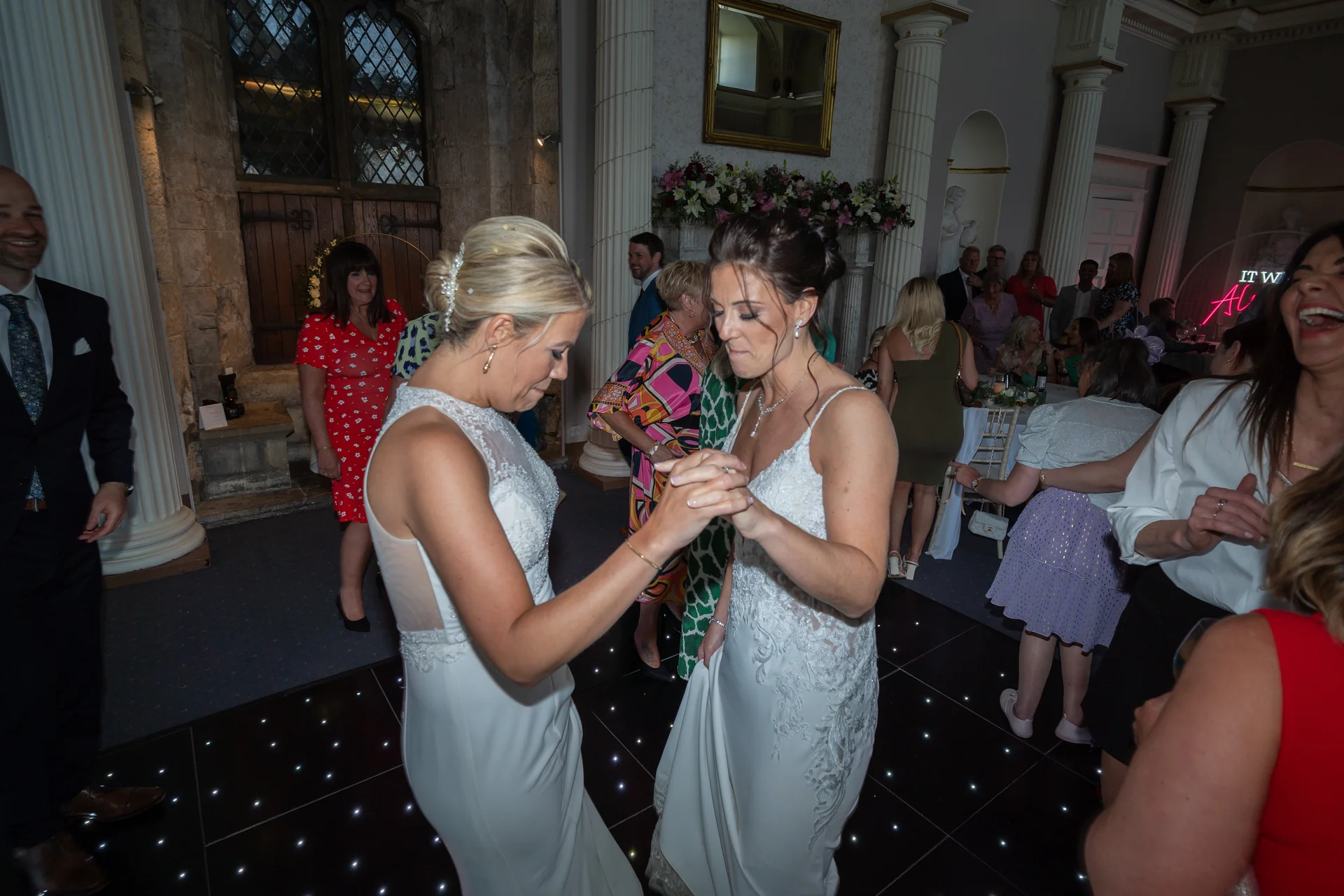 Two brides share their first dance together on a glowing dance floor surrounded by smiling family and guests inside a historic venue.