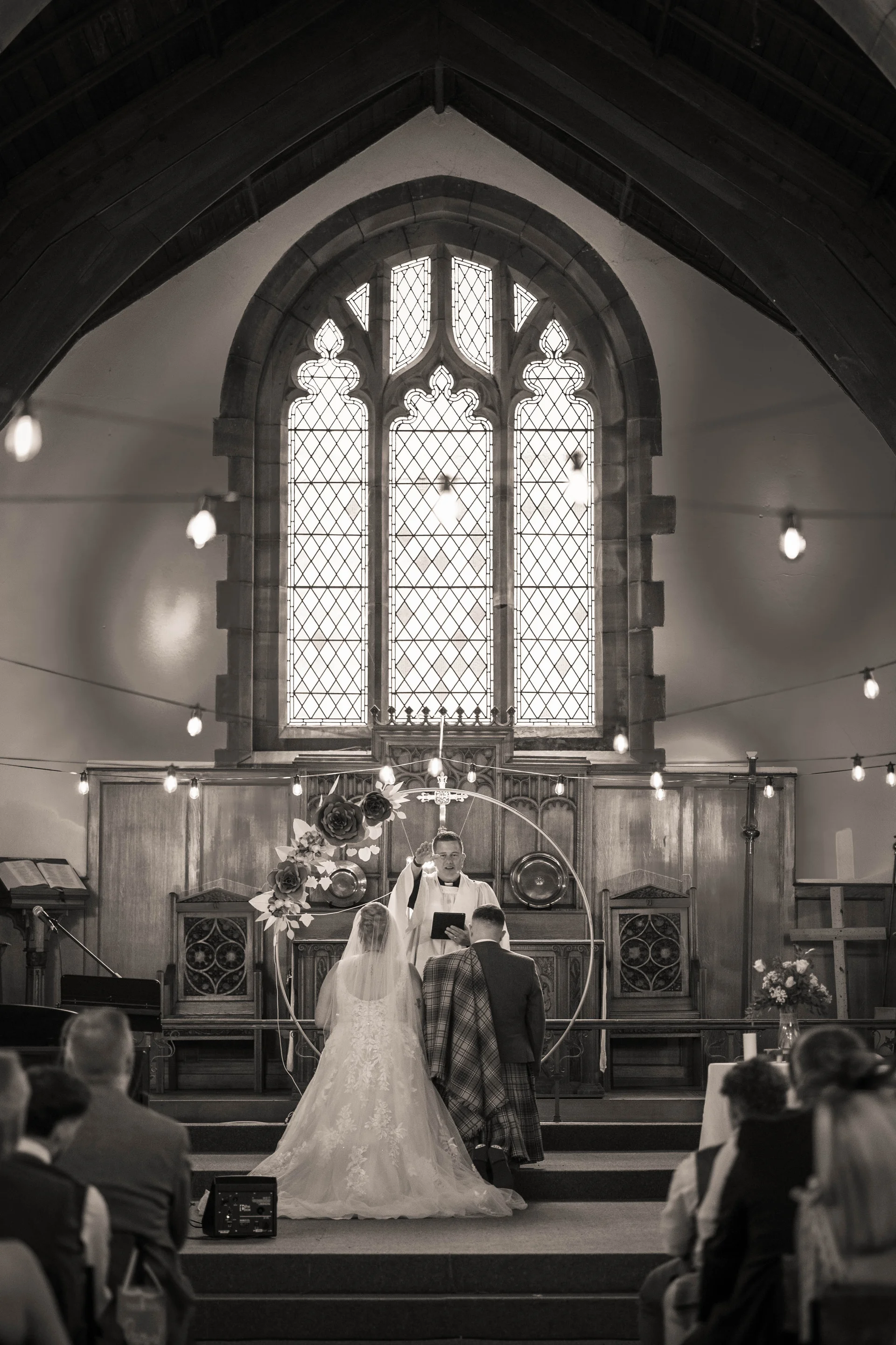A newlywed couple stands before an officiant at the altar of a historic church featuring a grand stained glass window and overhead string lighting.