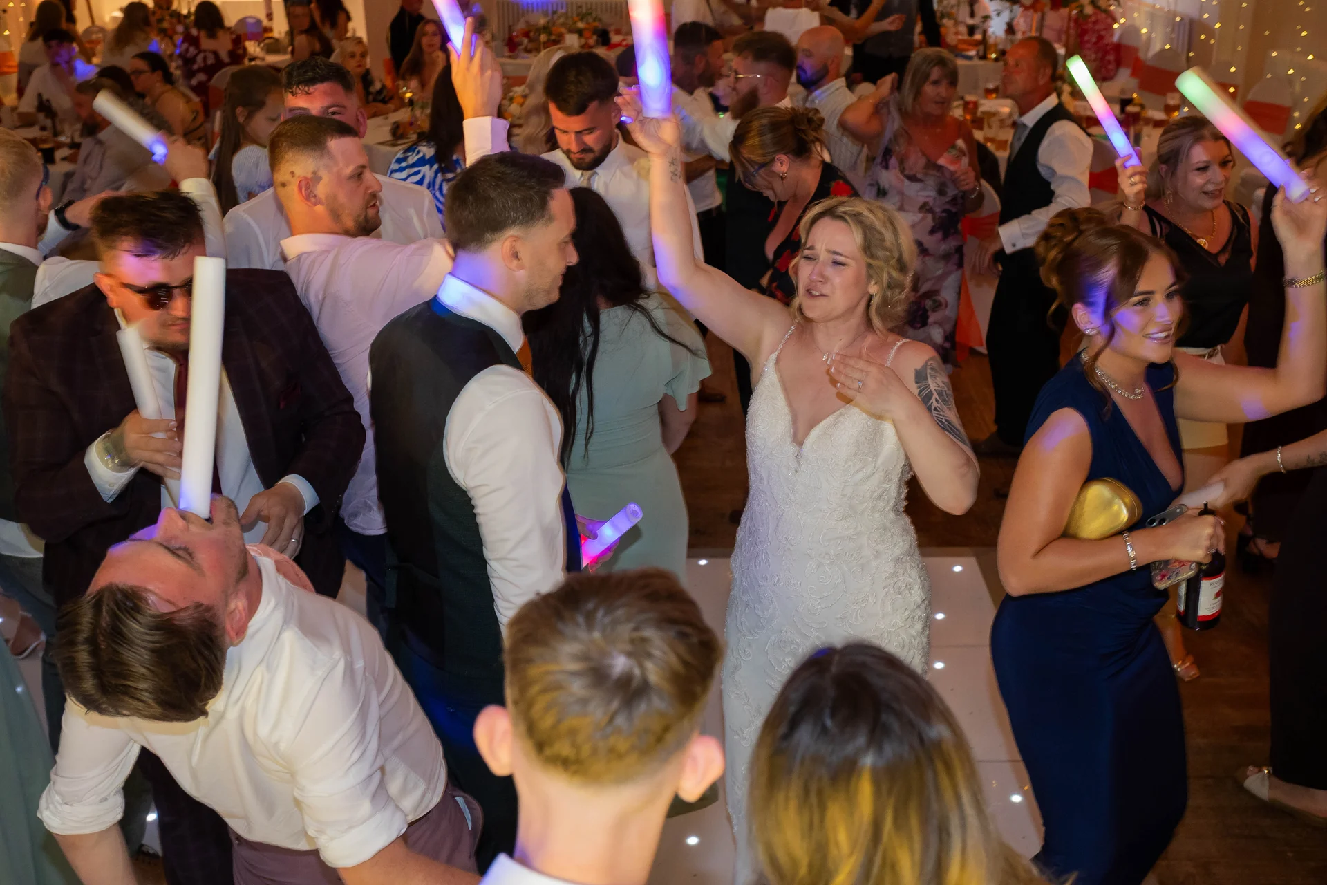 The bride and wedding guests celebrate enthusiastically on a glowing dance floor, waving light-up foam sticks amidst a lively party atmosphere.