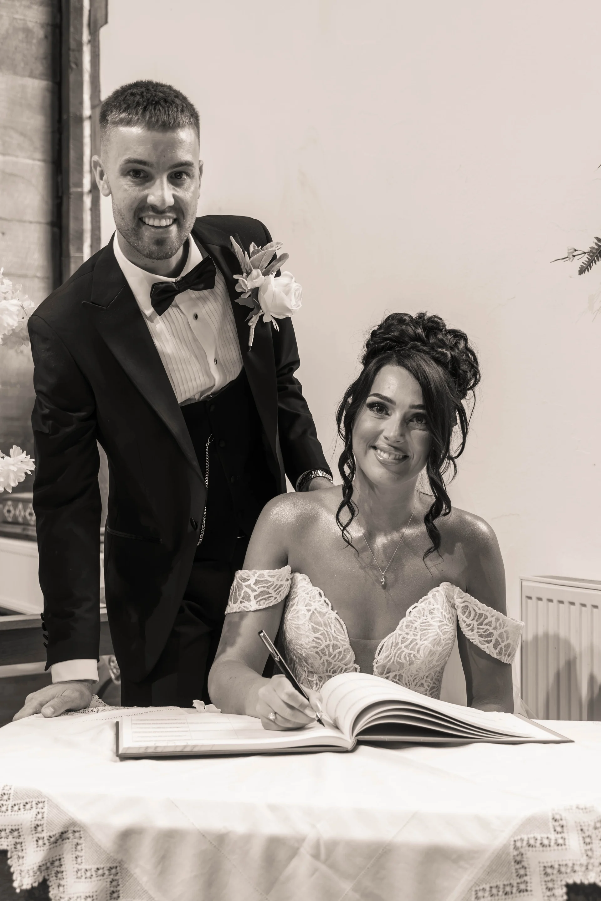 The bride and groom smile brightly as the bride signs the marriage register in sepia tones during their wedding ceremony.