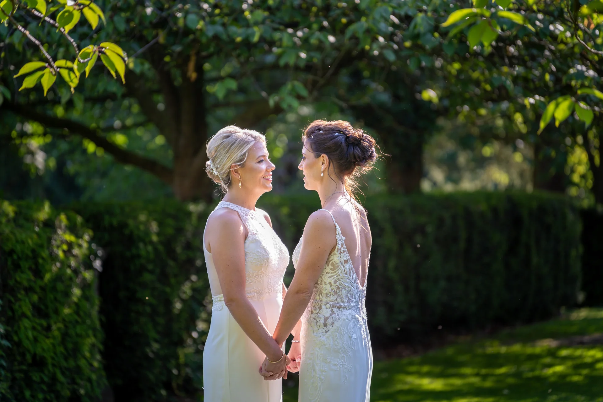 Two brides hold hands and look lovingly at each other in a sunlit garden during golden hour.