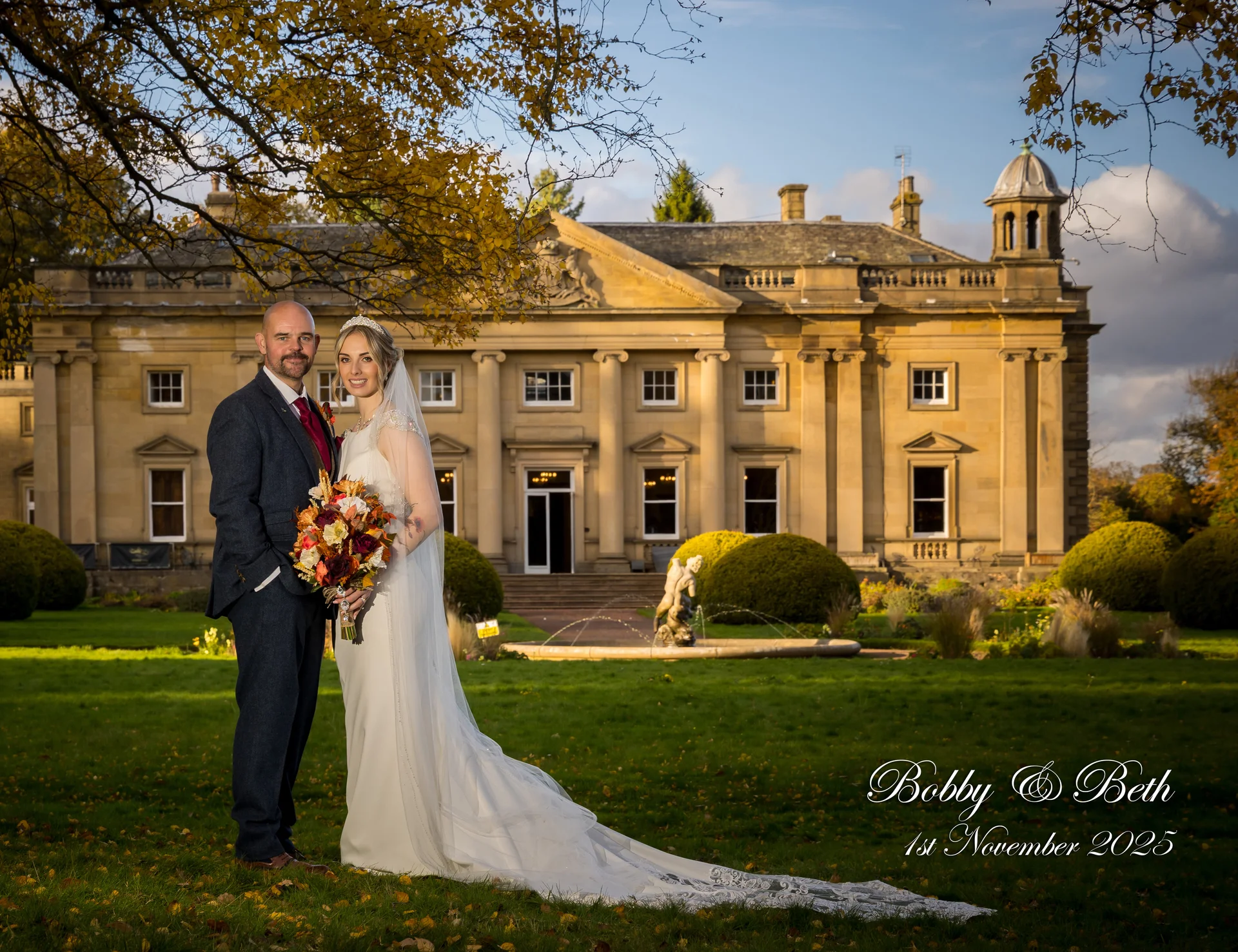 A bride and groom pose on the manicured lawn in front of a grand stone country house during an autumn wedding.