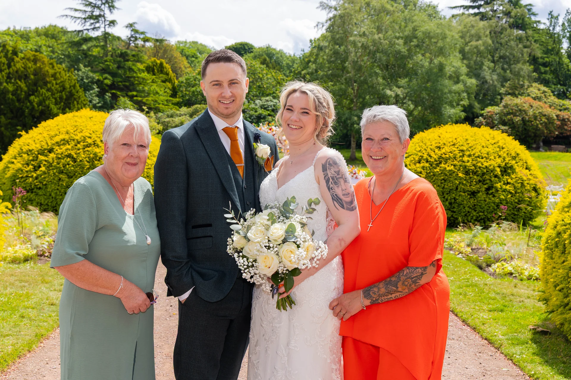 A smiling bride and groom pose outdoors in a sunny garden alongside two female family members, with the bride holding a white rose bouquet.