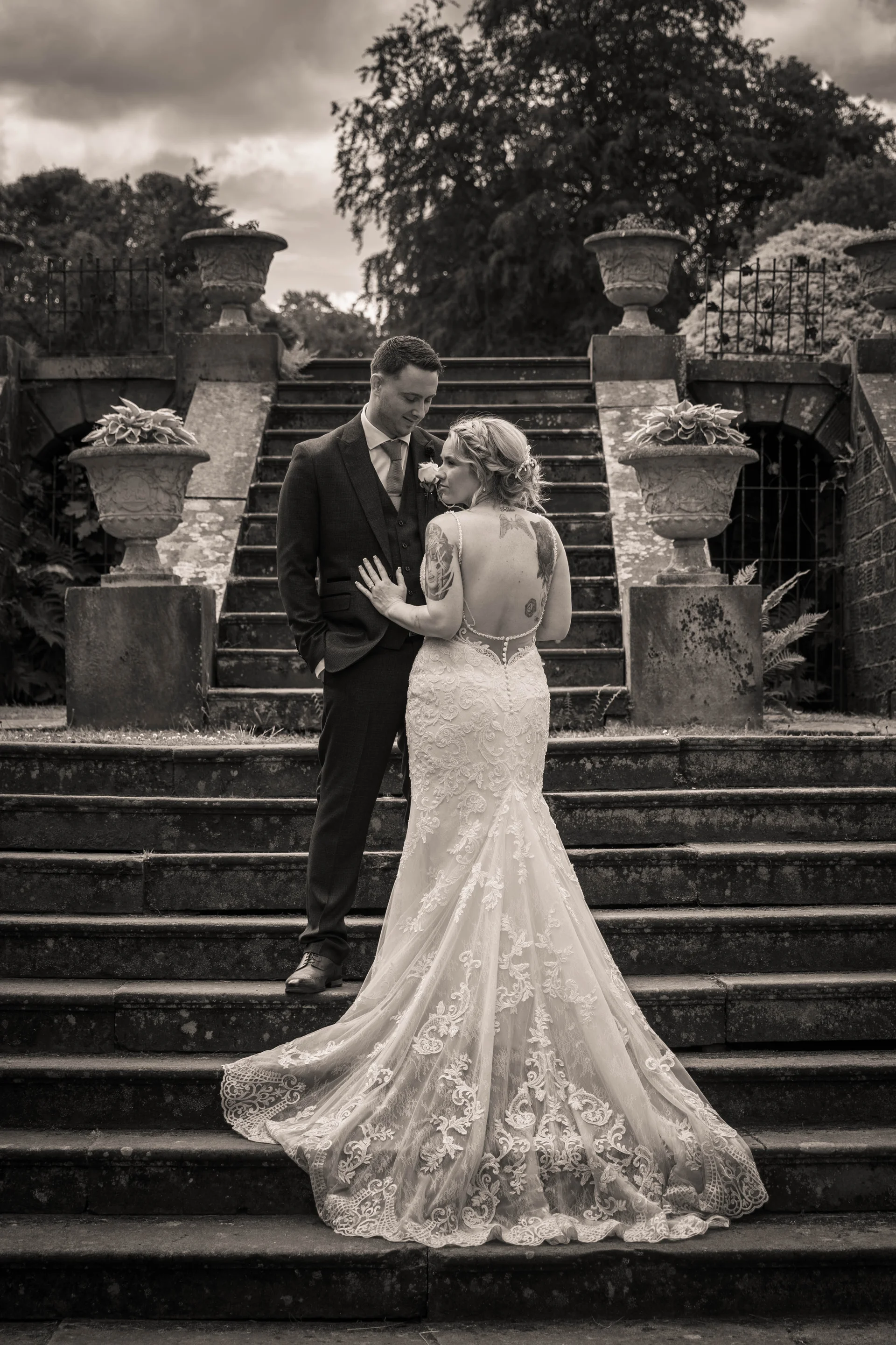 A newlywed couple poses on a grand stone staircase flanked by ornate urns, with the bride showcasing a detailed lace dress featuring an open back and tattoos.