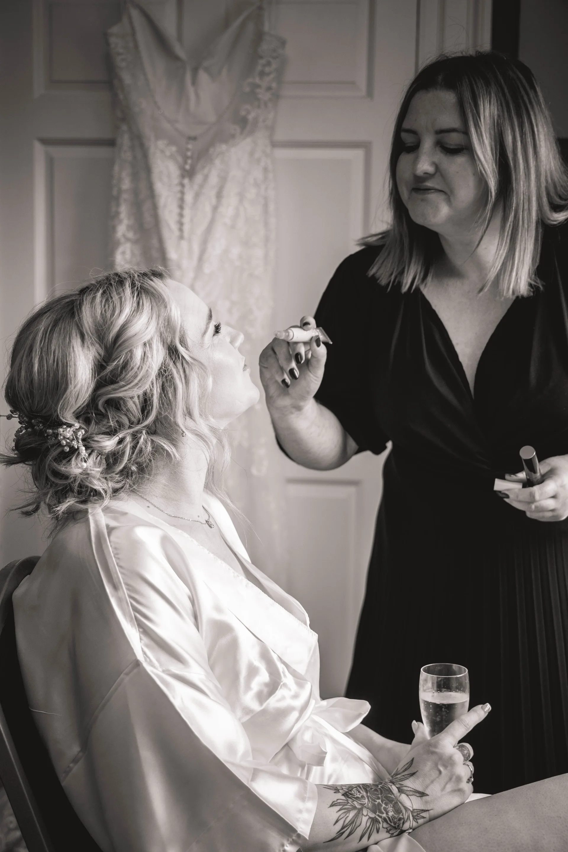 A black and white photograph captures a bride having her makeup applied while holding a glass of champagne, with her wedding dress hanging in the background.