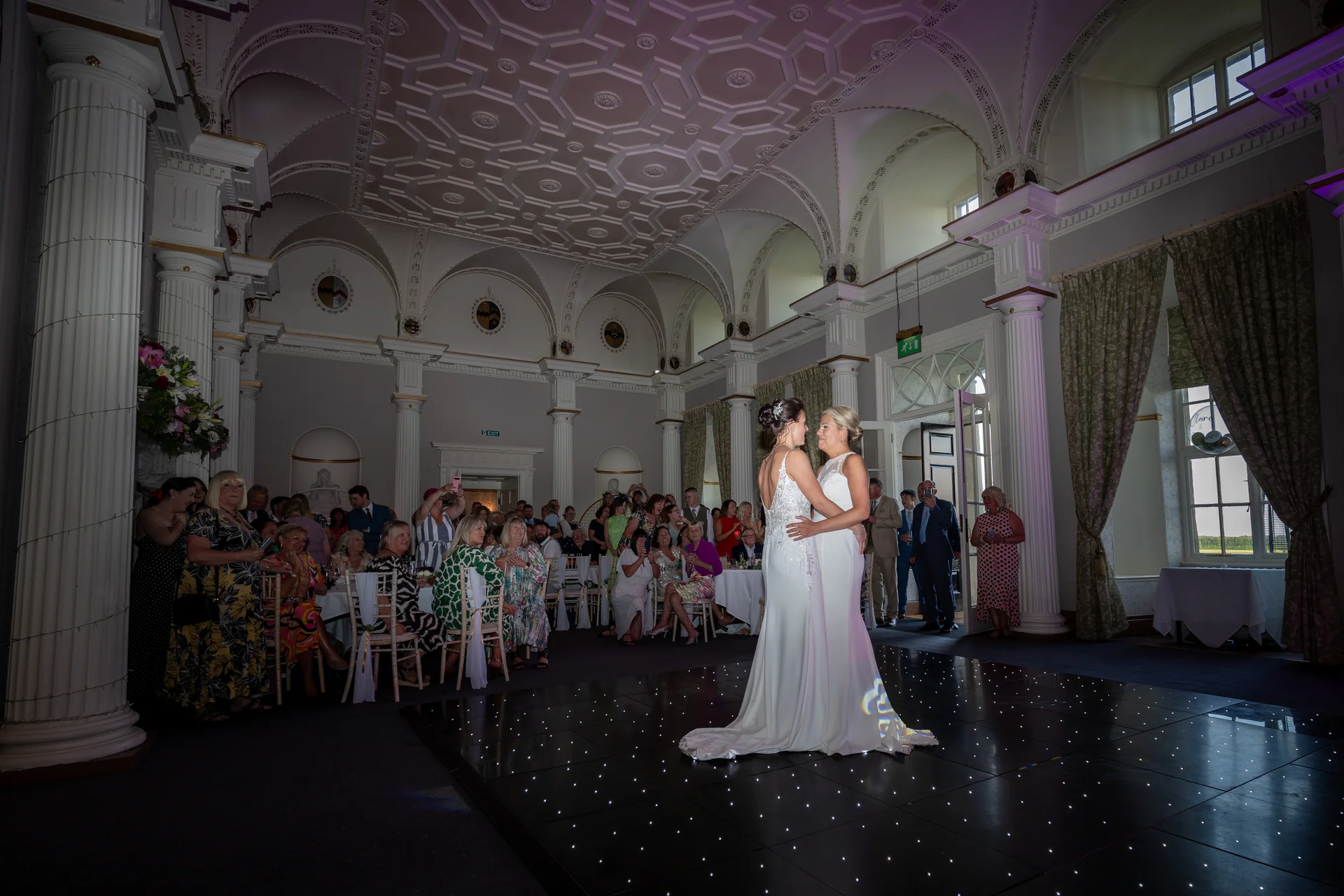 Two brides share a tender embrace during their first dance on a starlit dance floor inside a grand stately home ballroom filled with seated guests.