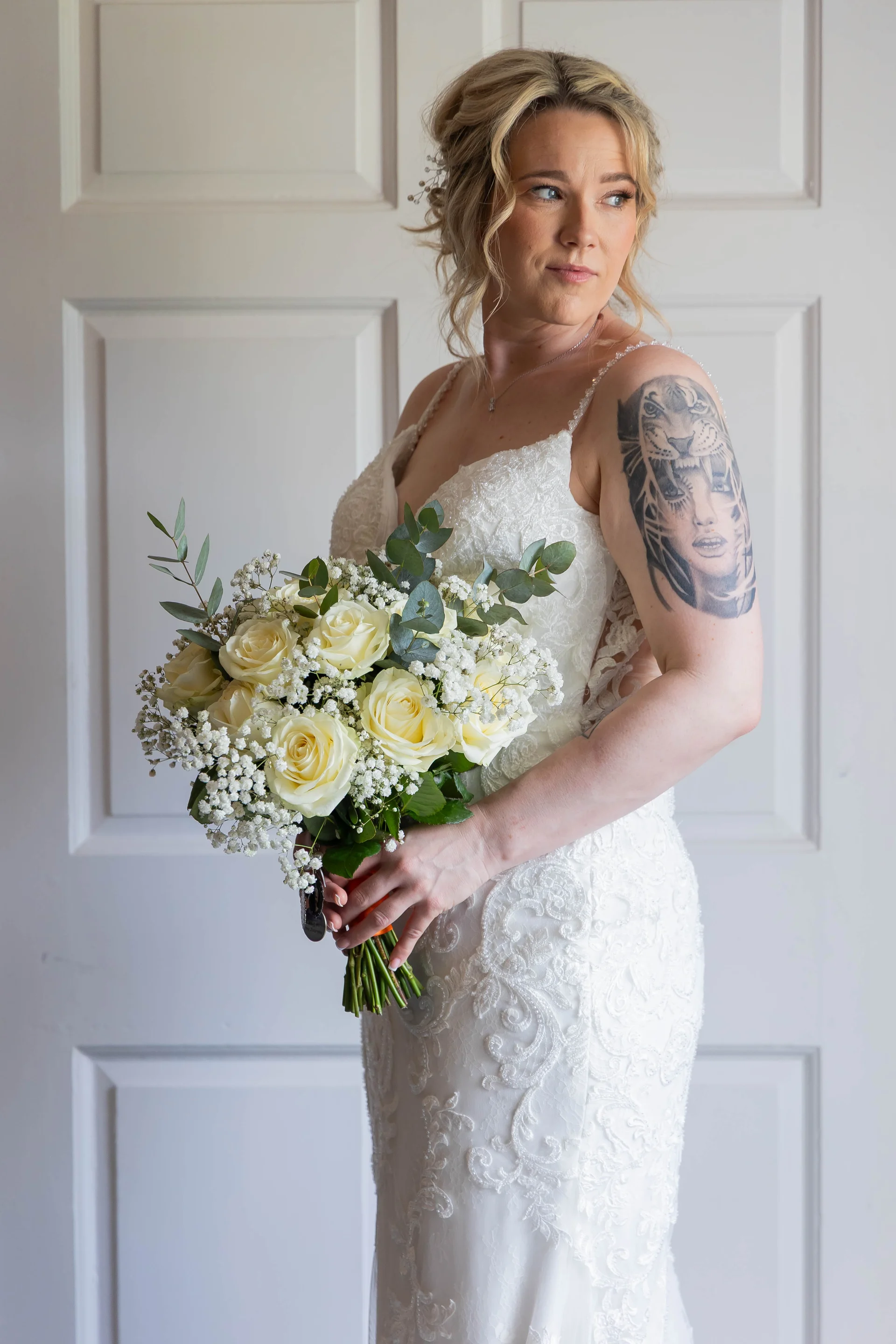 A stunning portrait of a bride wearing a lace beaded gown with spaghetti straps, holding a hand-tied bouquet of white roses and baby's breath, showcasing a large arm tattoo.