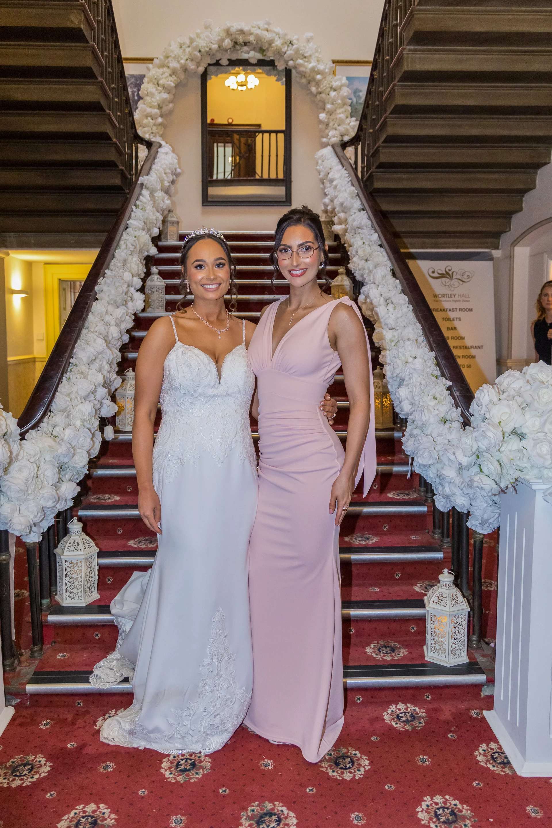 Two brides pose together smiling on a grand carpeted staircase adorned with cascading white rose garlands inside a stately home.