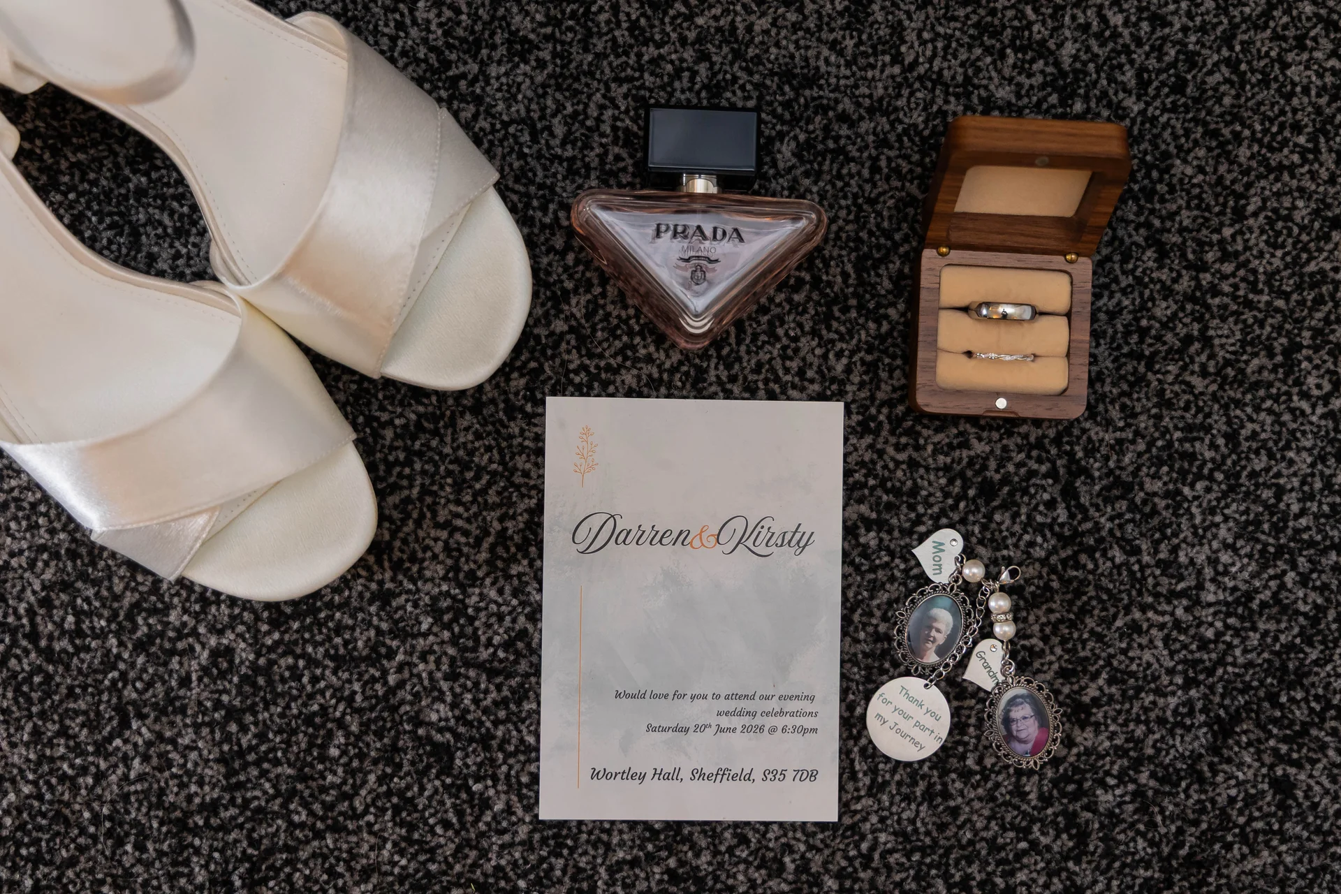 A flat lay image of bridal accessories including white satin heels, a wedding invitation for Darren and Kirsty, a Prada perfume bottle, wedding bands in a wooden ring box, and commemorative photo charms resting on dark textured carpet.
