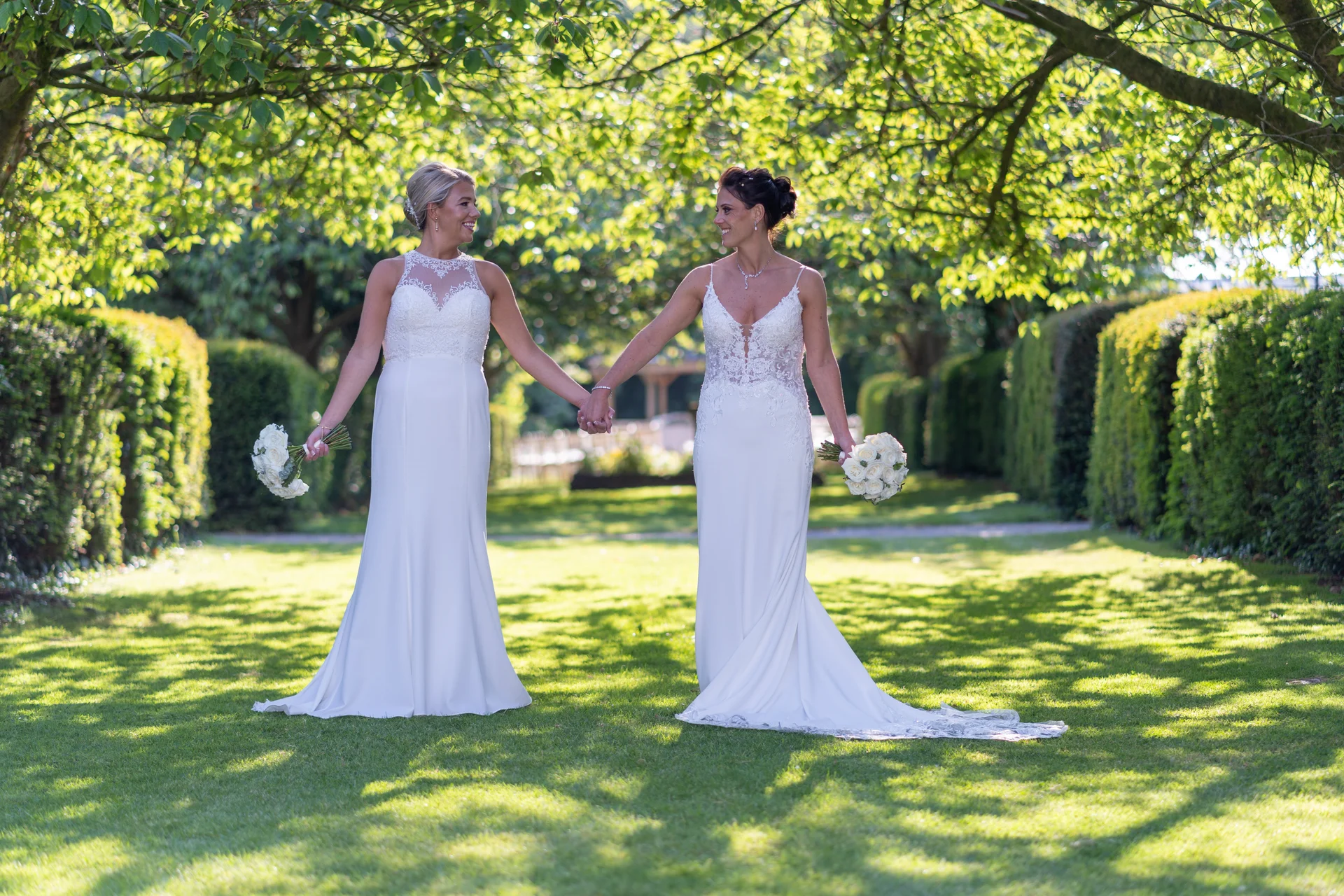 Two smiling brides in white wedding dresses walk hand in hand through a sunlit, tree-lined garden path, each holding a bouquet of white roses.
