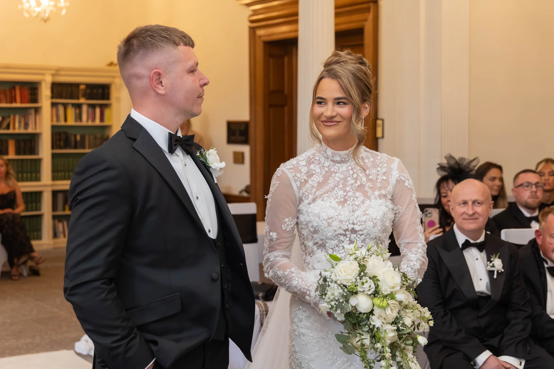 A smiling bride holding a white floral bouquet looks toward the groom at the altar during their ceremony inside a historic library venue.