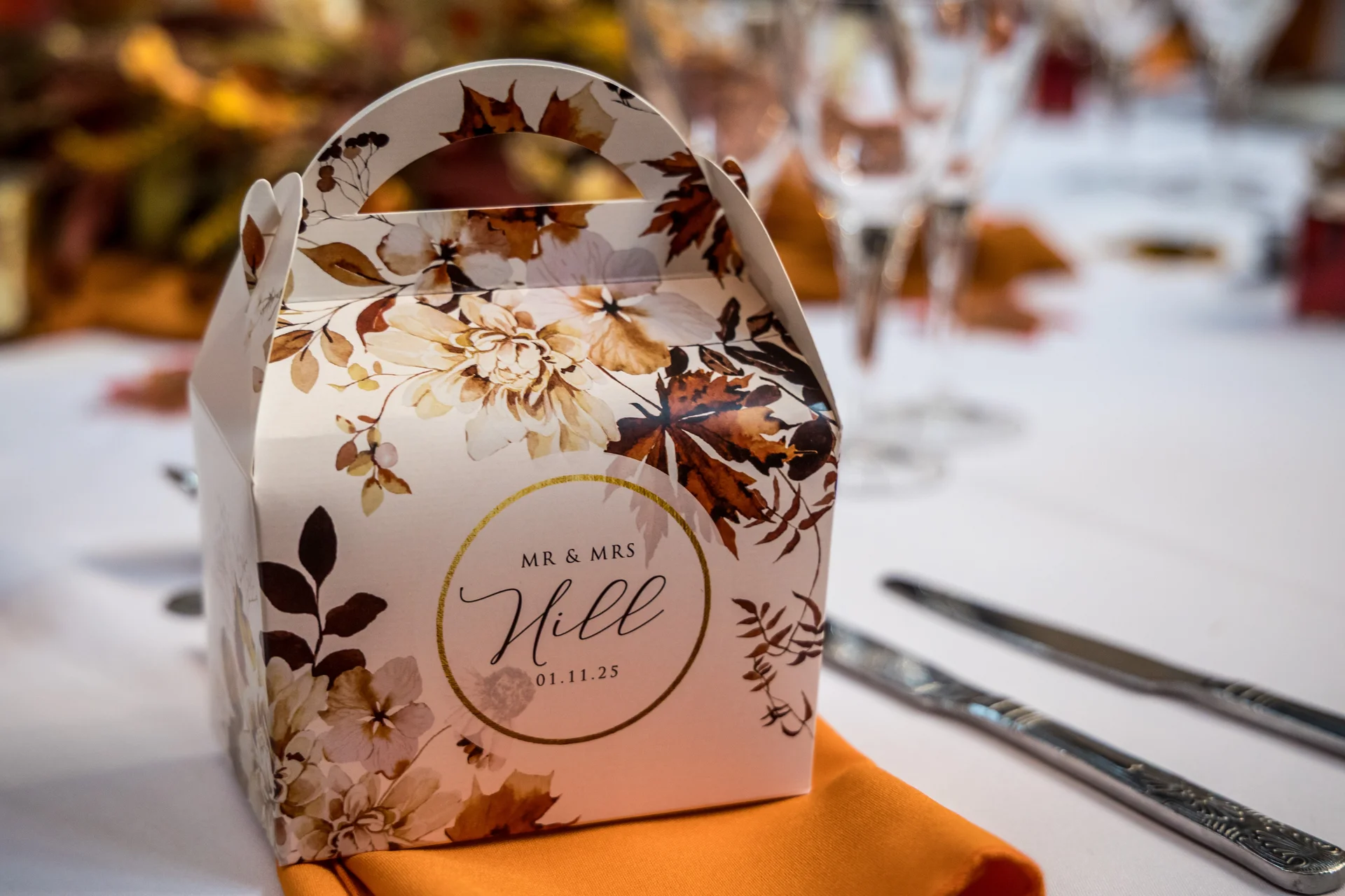 A personalised autumn-themed favour box featuring floral artwork and the couple's name and wedding date rests on an orange napkin at a wedding reception.