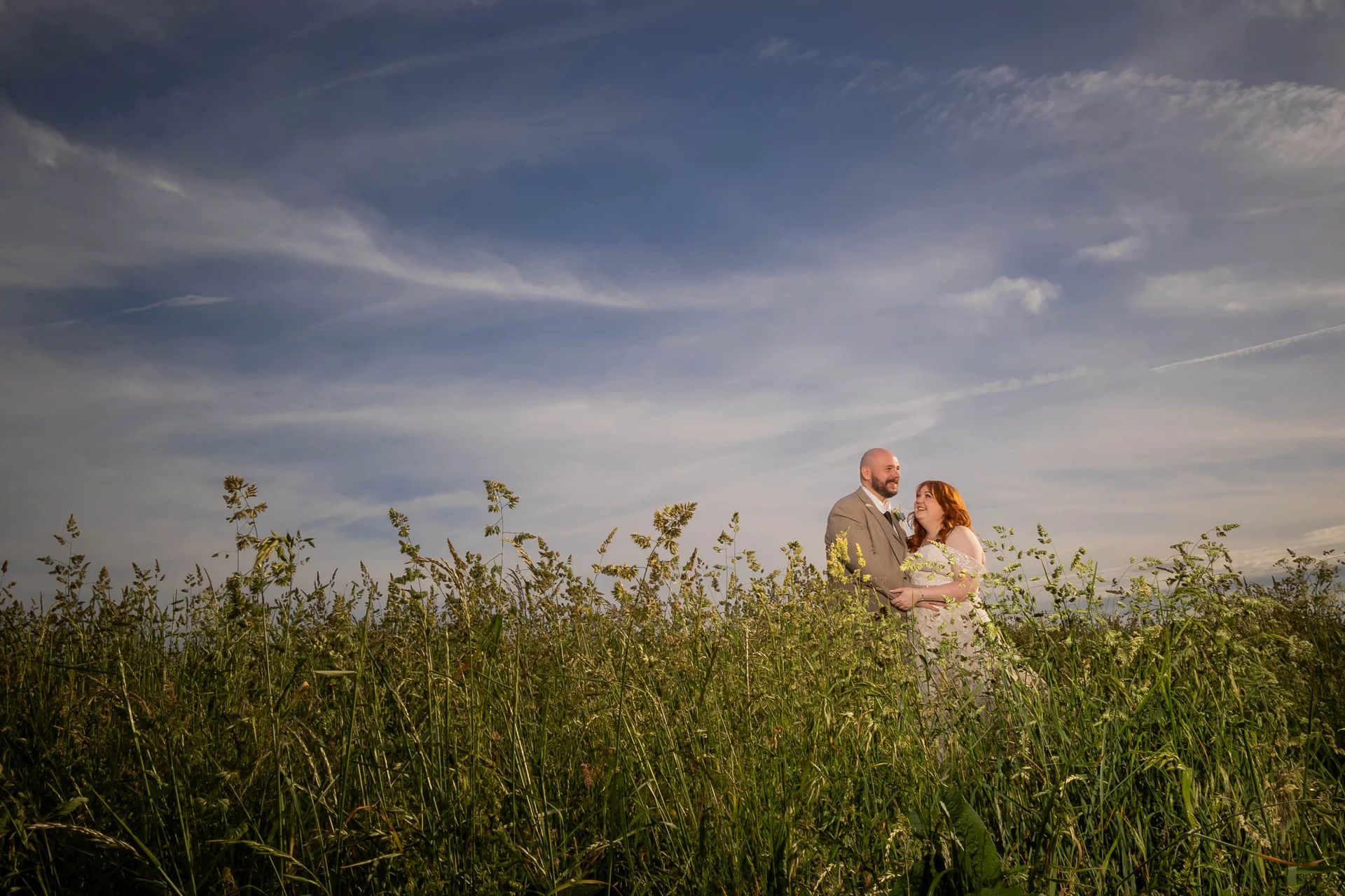 A bride with vibrant red hair and a groom stand embraced in a tall wildflower meadow beneath a vast, sweeping blue sky during golden hour.