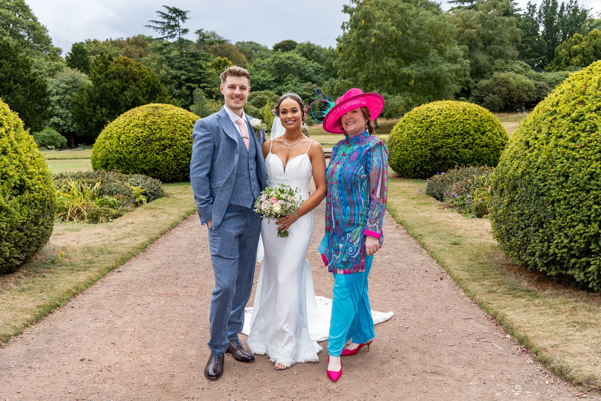 A smiling bride and groom pose on a gravel pathway in a manicured English country garden alongside a guest wearing a vibrant pink hat and patterned tunic.