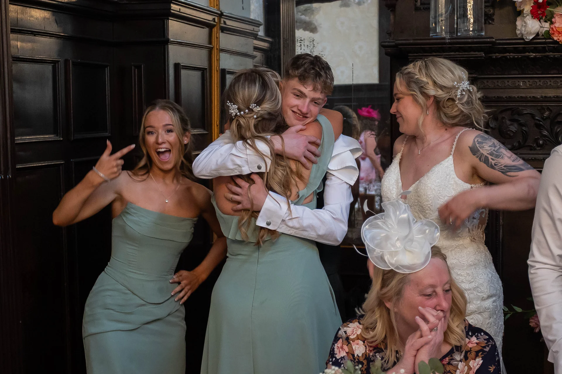A bride in a lace gown and a bridesmaid look on smiling as a couple embraces warmly, while another bridesmaid pulls a joyful face.