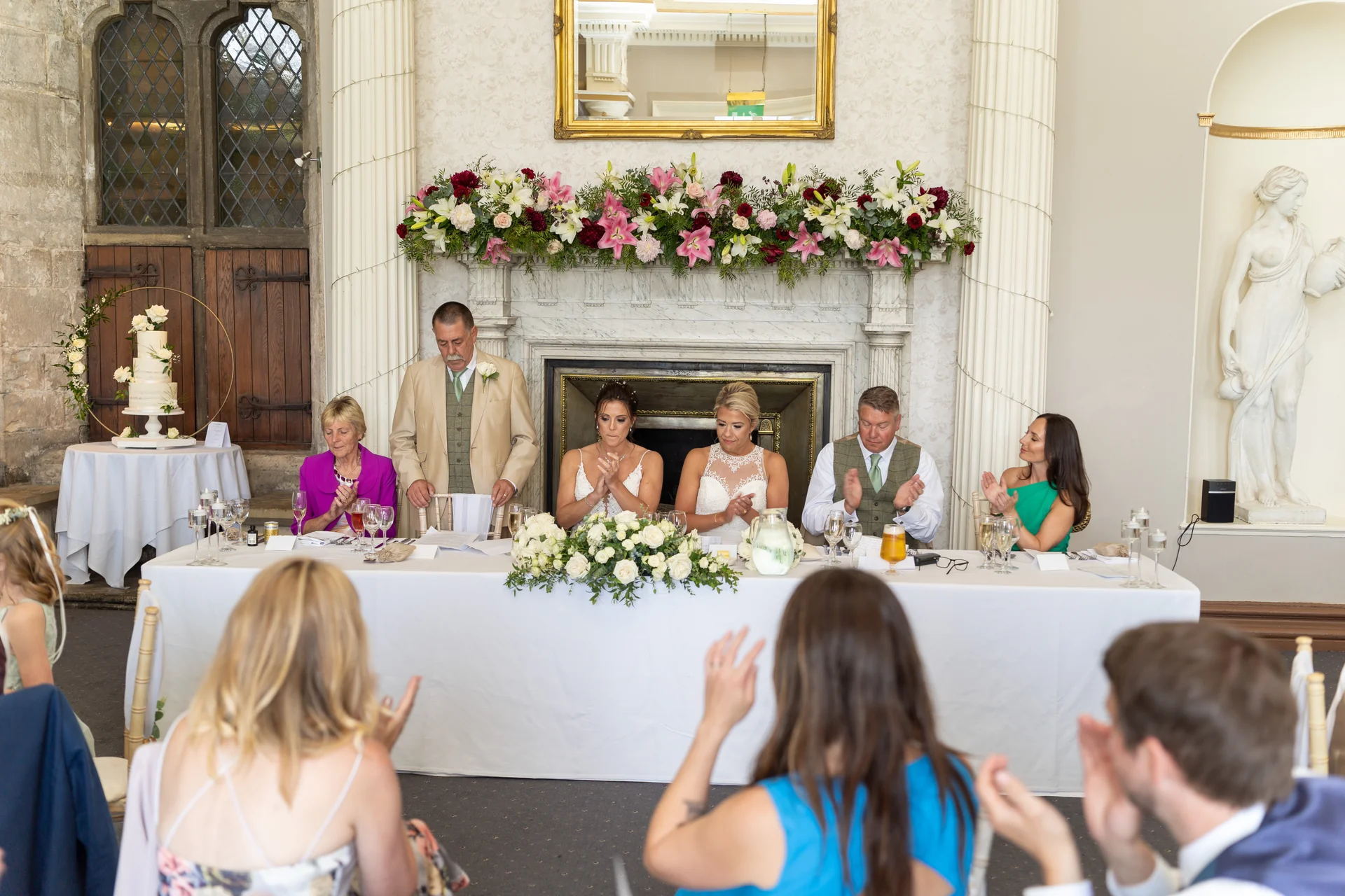 The wedding party and guests applaud at the top table during speeches in a grand country house reception room.
