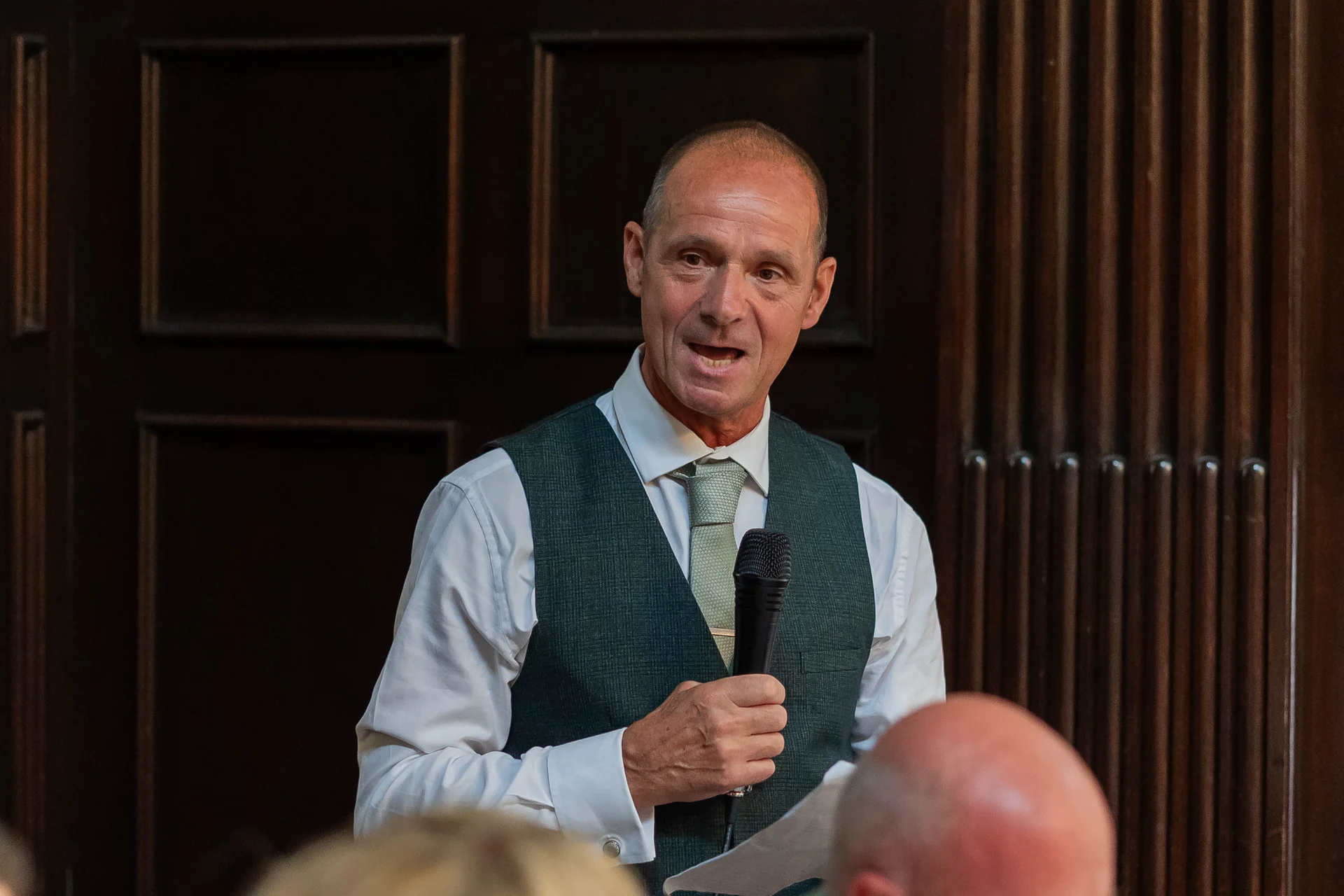 A mature groom wearing an olive green waistcoat and patterned tie delivers a speech into a microphone during the wedding reception.