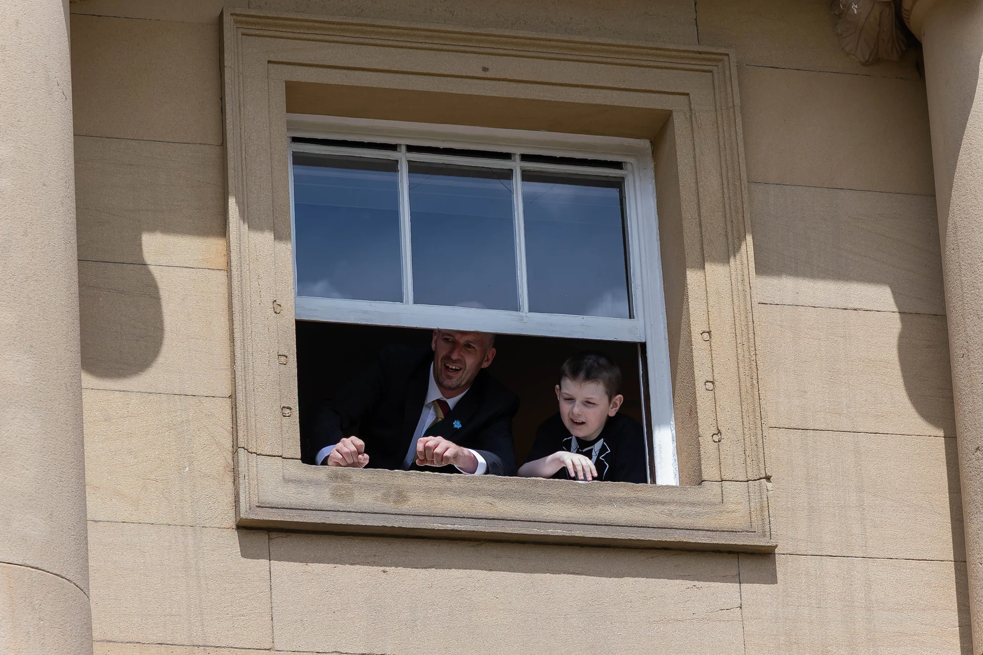 A man and a young boy lean out of a stone window on a stately home, laughing and looking down at the wedding celebrations below.