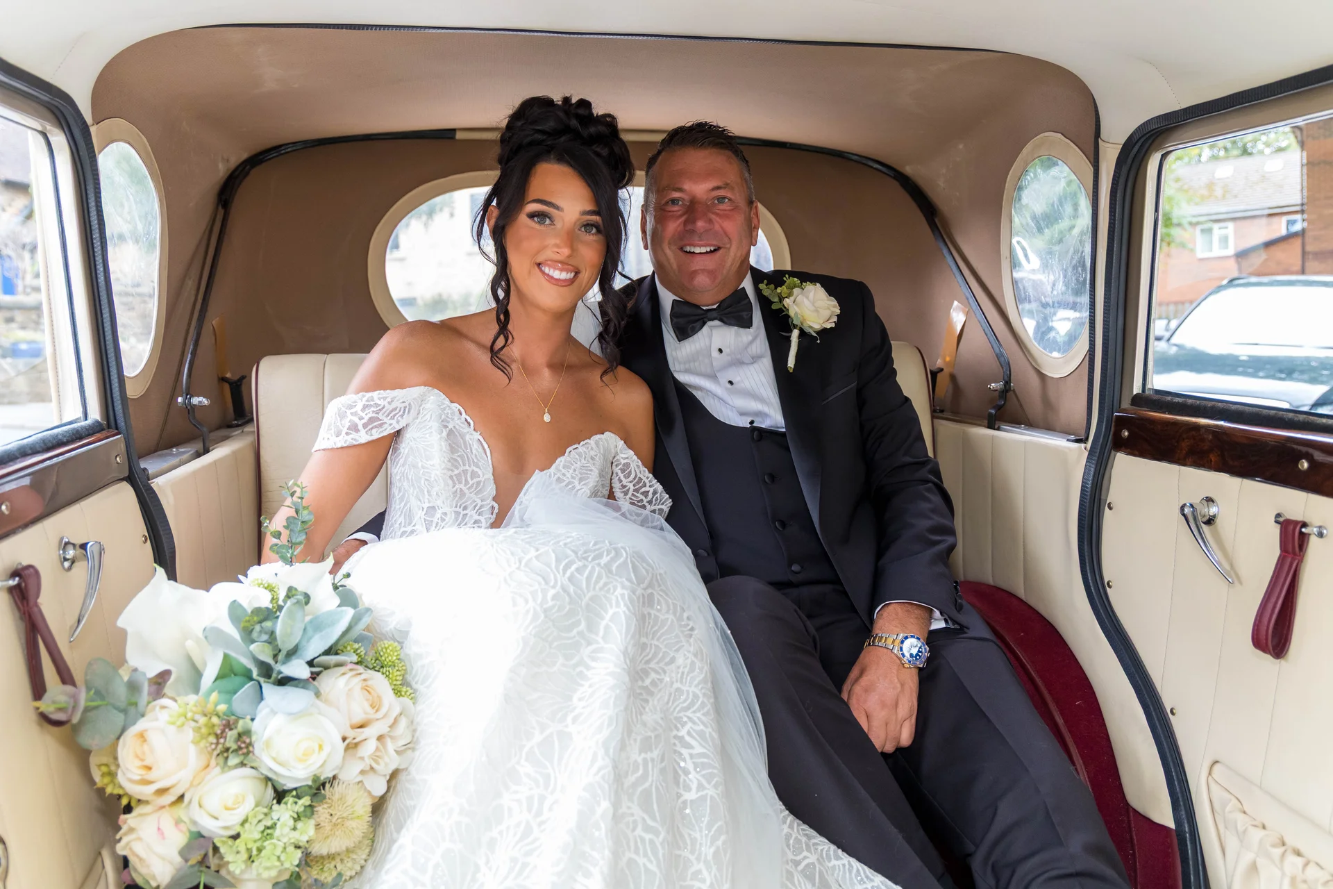 A smiling bride and her father sit inside the luxurious interior of a classic vintage wedding car, with the bride holding a large white rose bouquet.