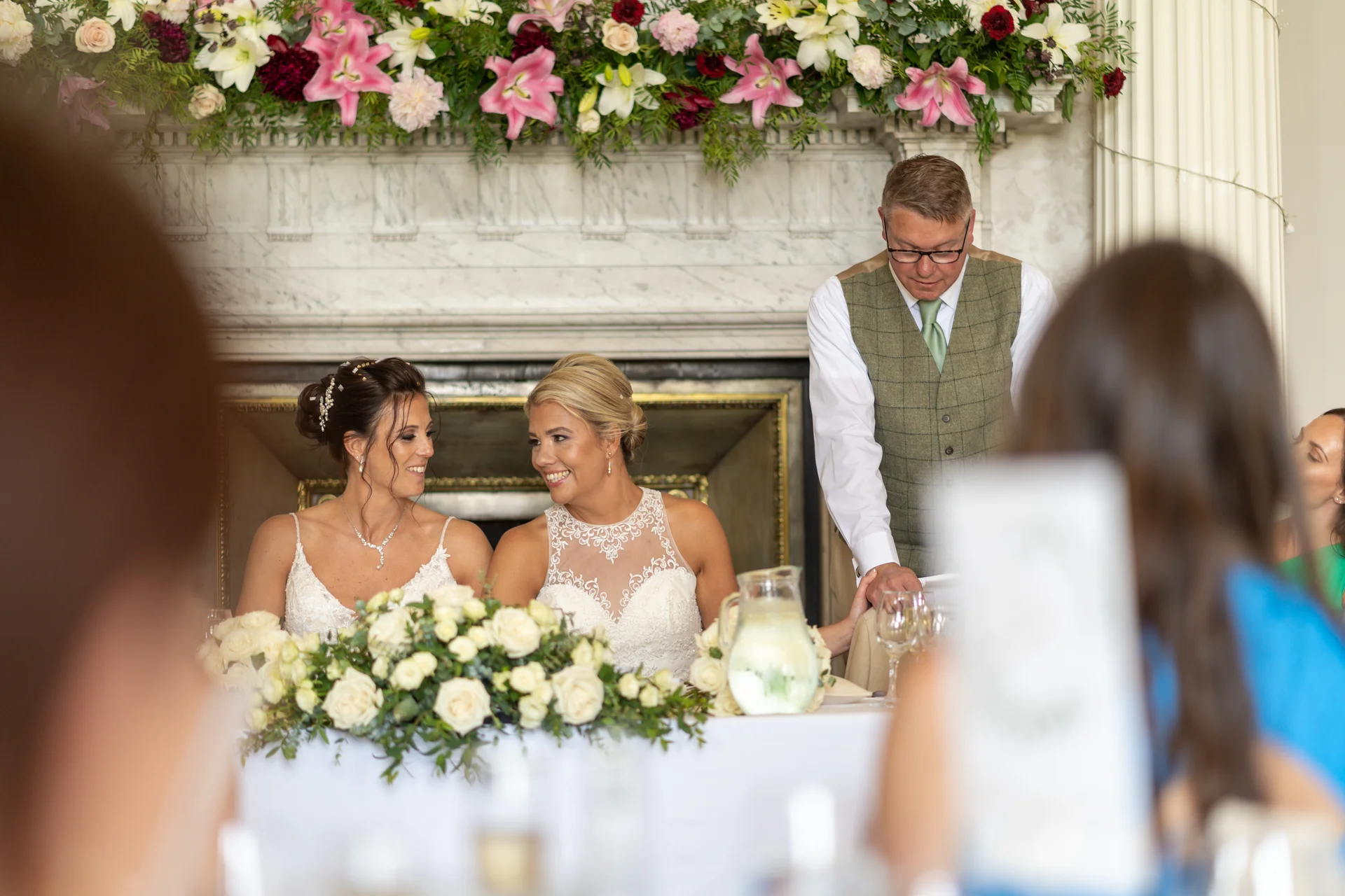 Two smiling brides sit together at the top table during wedding speeches, framed by a magnificent floral arrangement on the mantelpiece behind them.