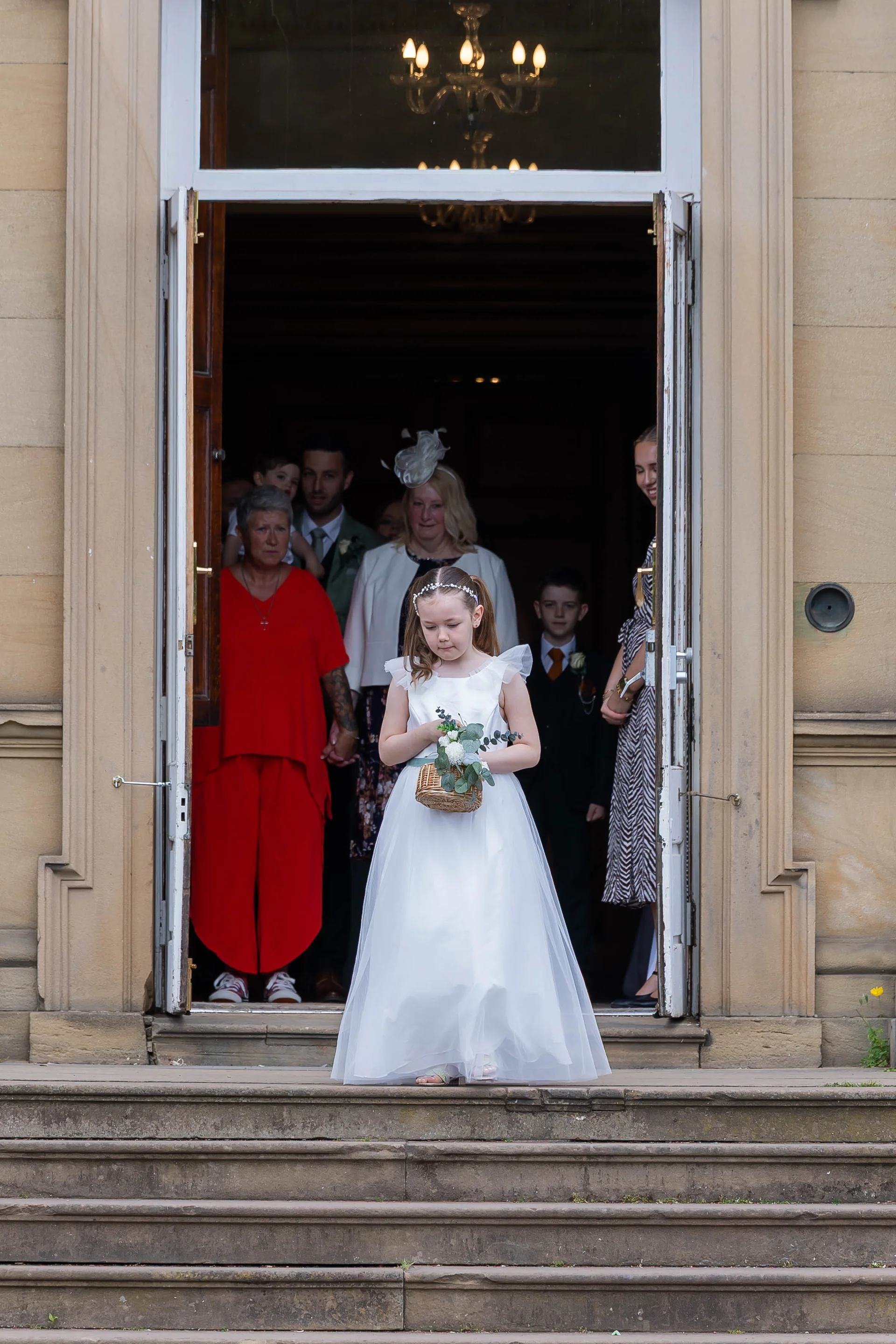 A young flower girl in a white dress carrying a small floral basket walks down stone steps from a country house doorway as wedding guests look on from inside.