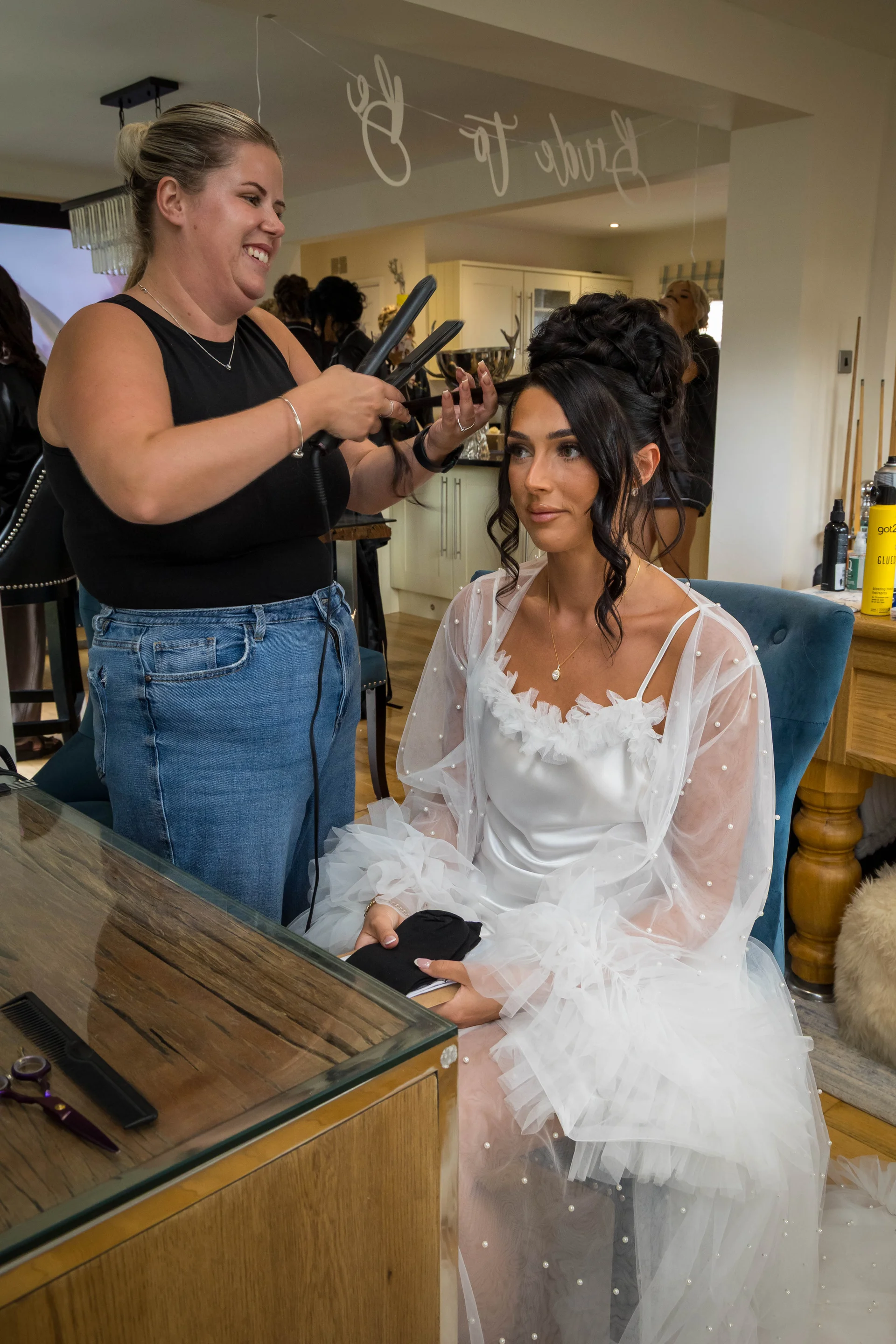 A smiling bride sits in a blue armchair while a hairstylist styles her hair with straighteners during bridal morning preparations.