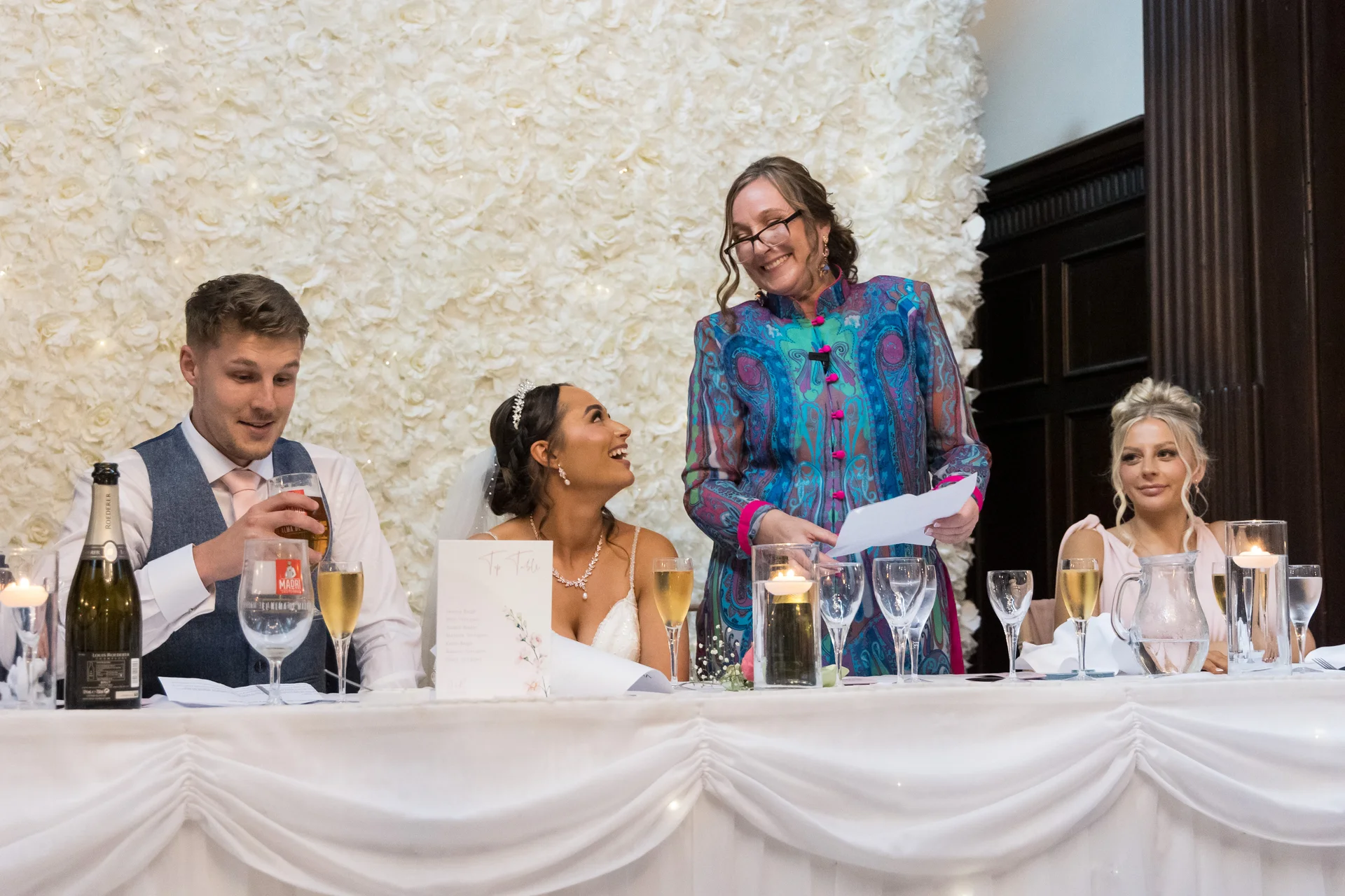 A smiling bride and groom sit at the top table sharing a lighthearted moment while a family member delivers a speech in front of a white floral wall.