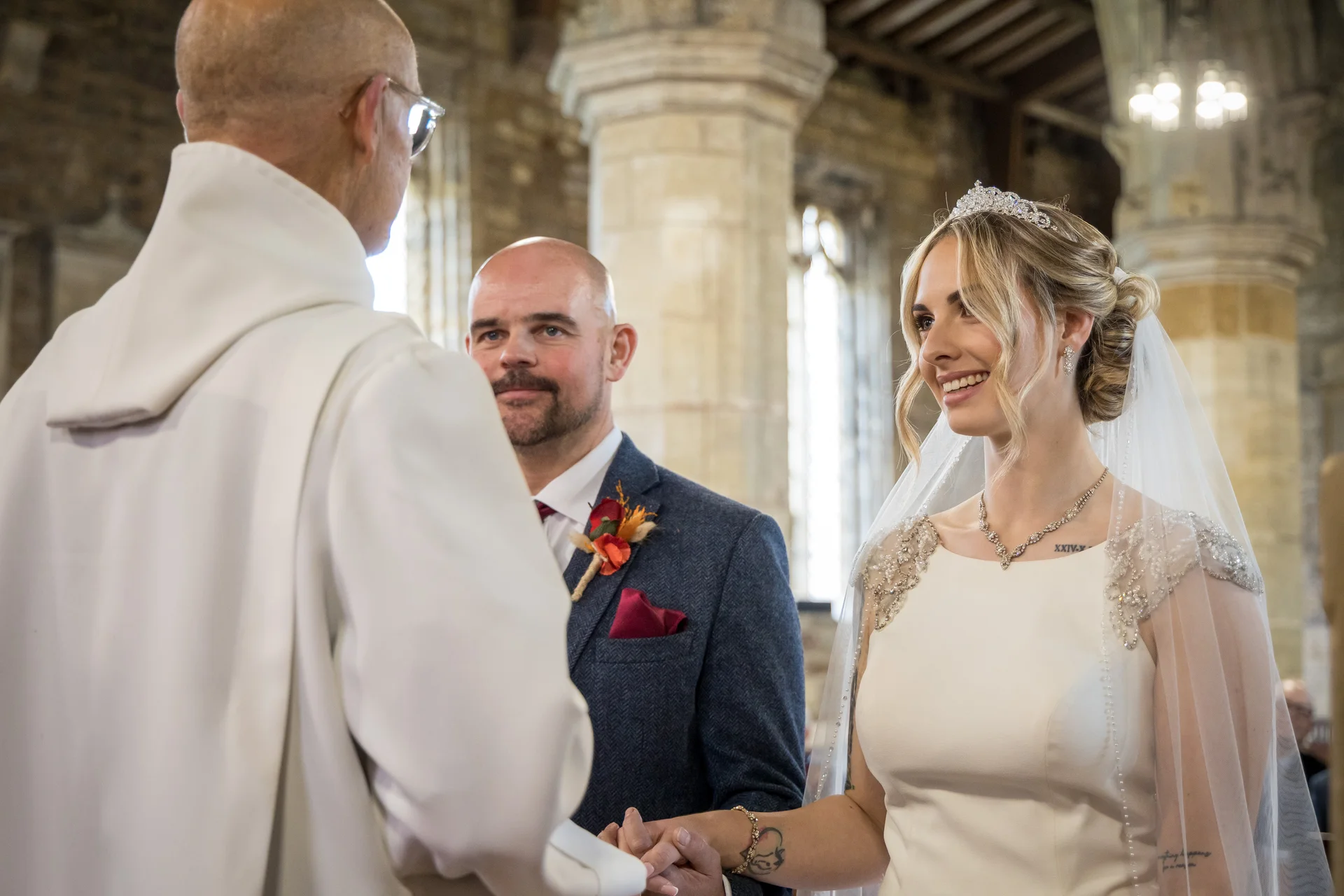 A smiling bride holding hands with her groom during a church wedding ceremony, standing before a robed officiant inside a historic stone building.