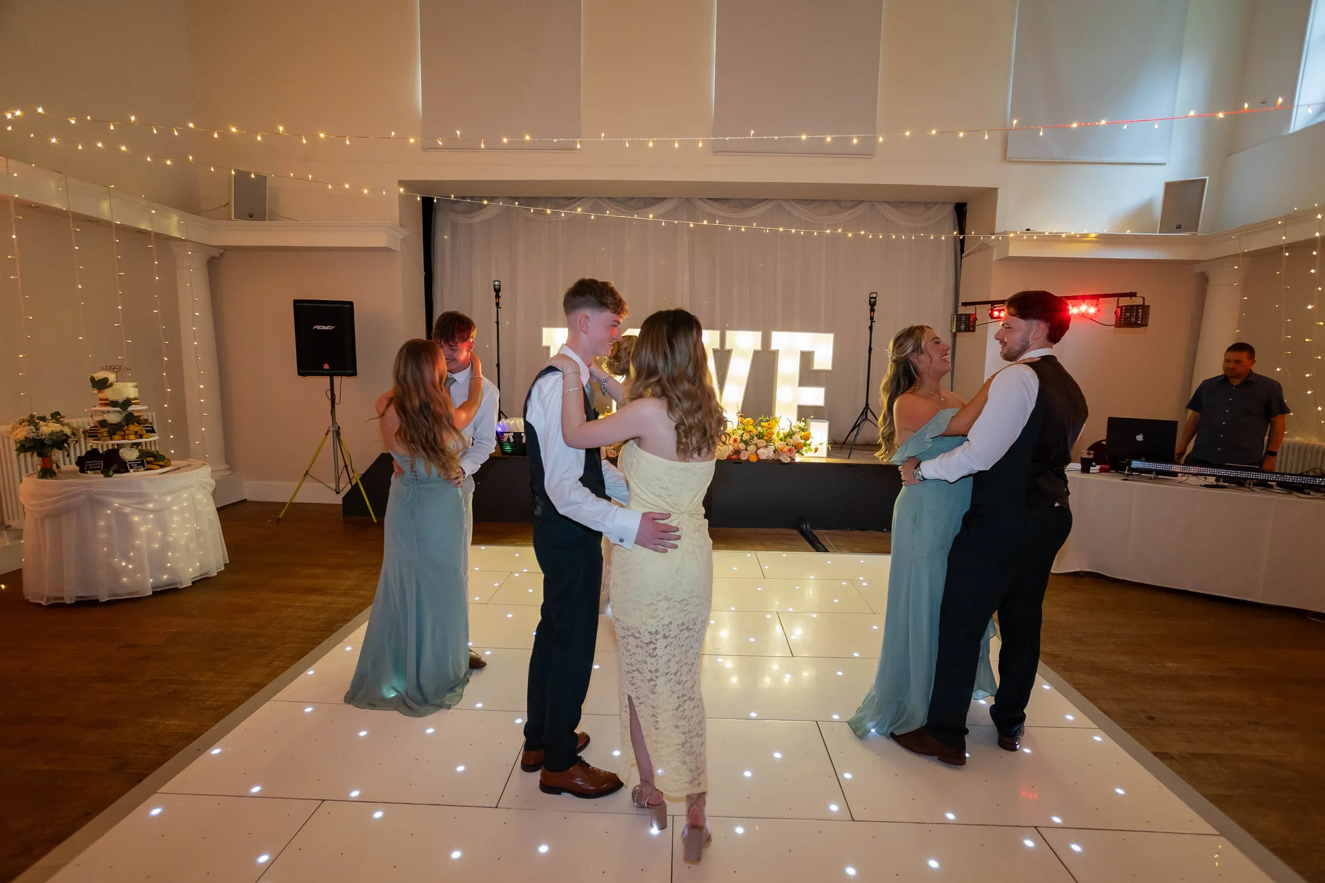 Several couples dance happily together on a lighted white dance floor under a canopy of delicate string lights in a spacious wedding venue hall.
