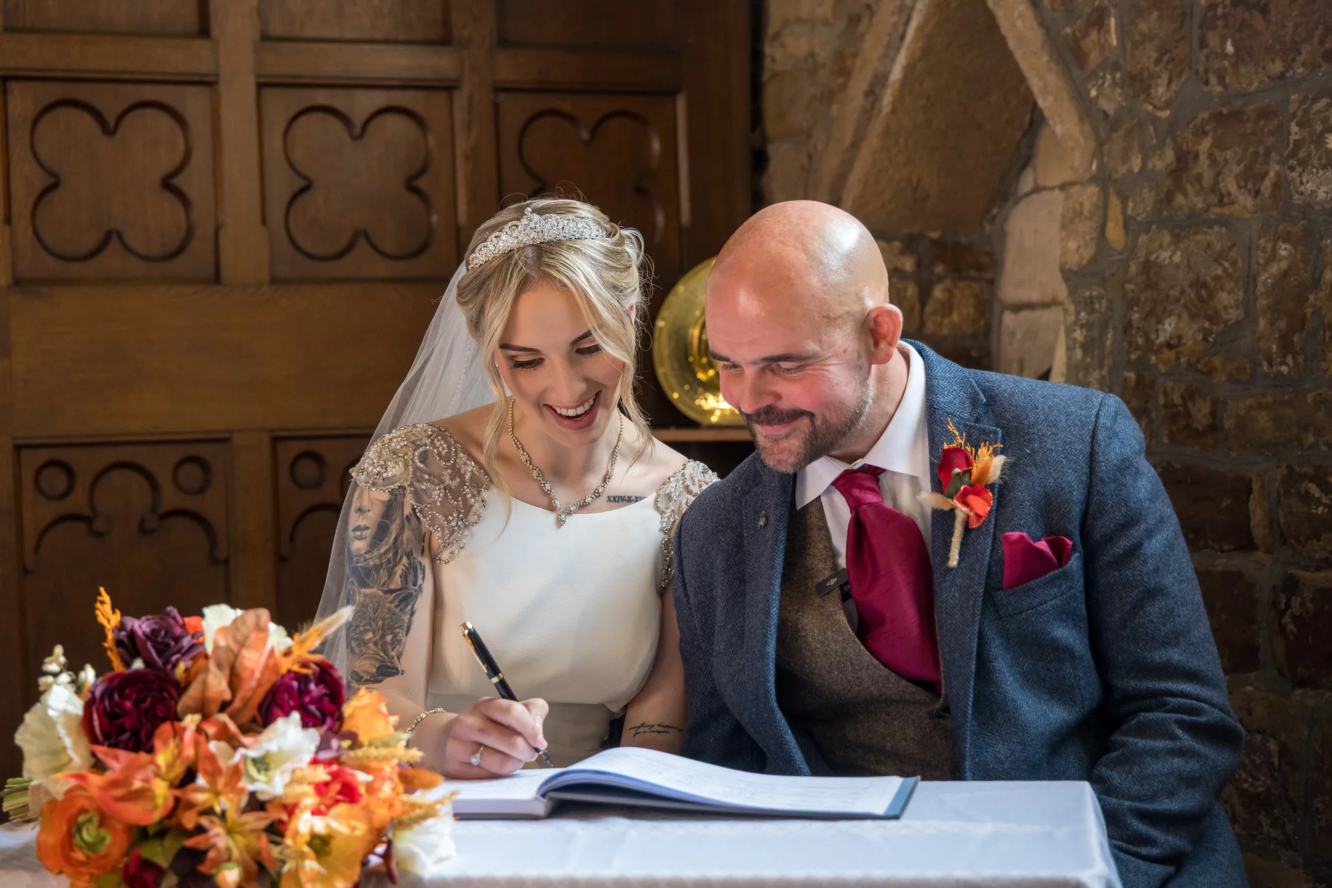 A smiling bride with tattoos and a crystal tiara signs the wedding register alongside her groom, who wears a tweed suit and a red tie.