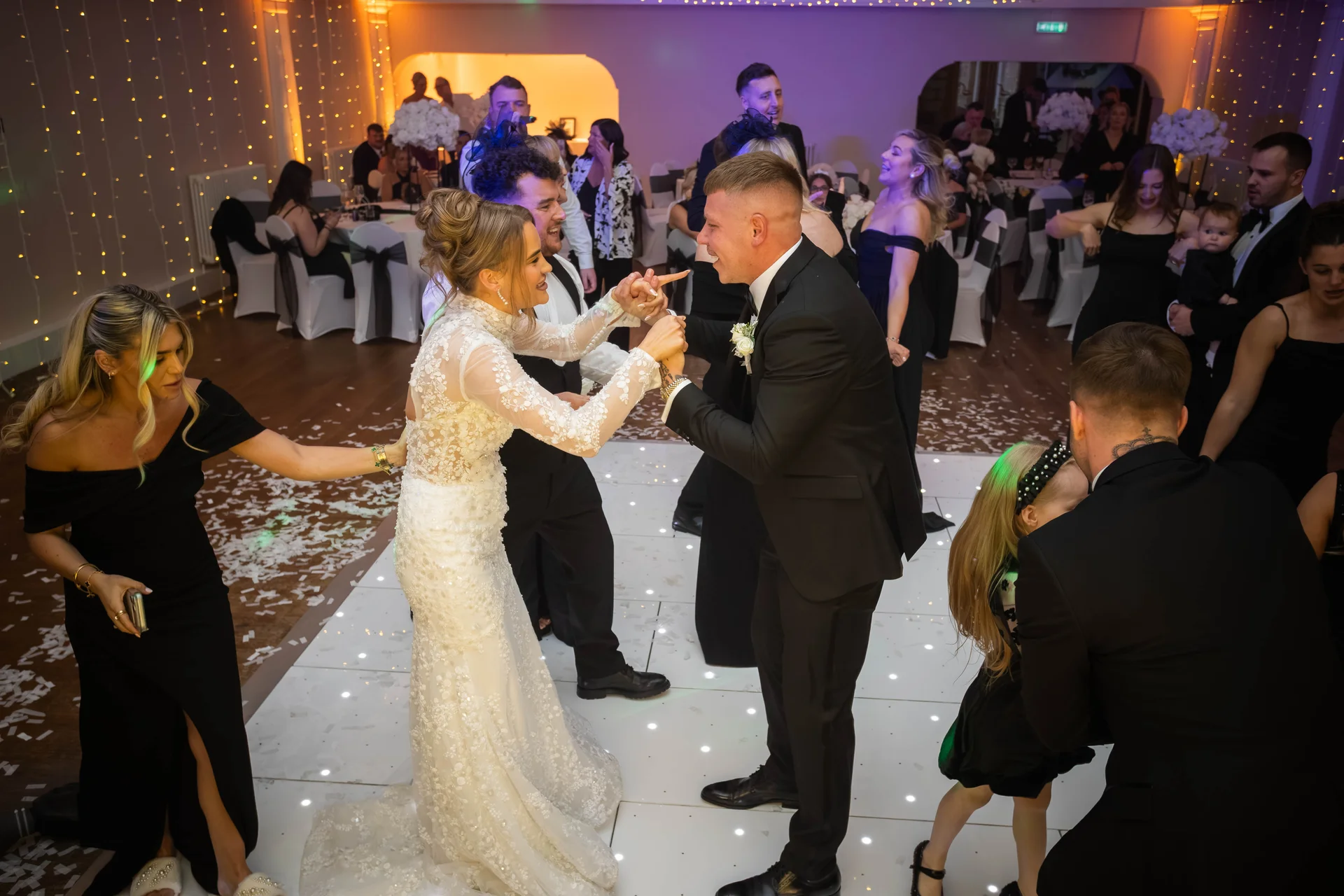 The newlywed couple joyfully share their first dance on a lit dance floor surrounded by wedding guests at an evening reception.