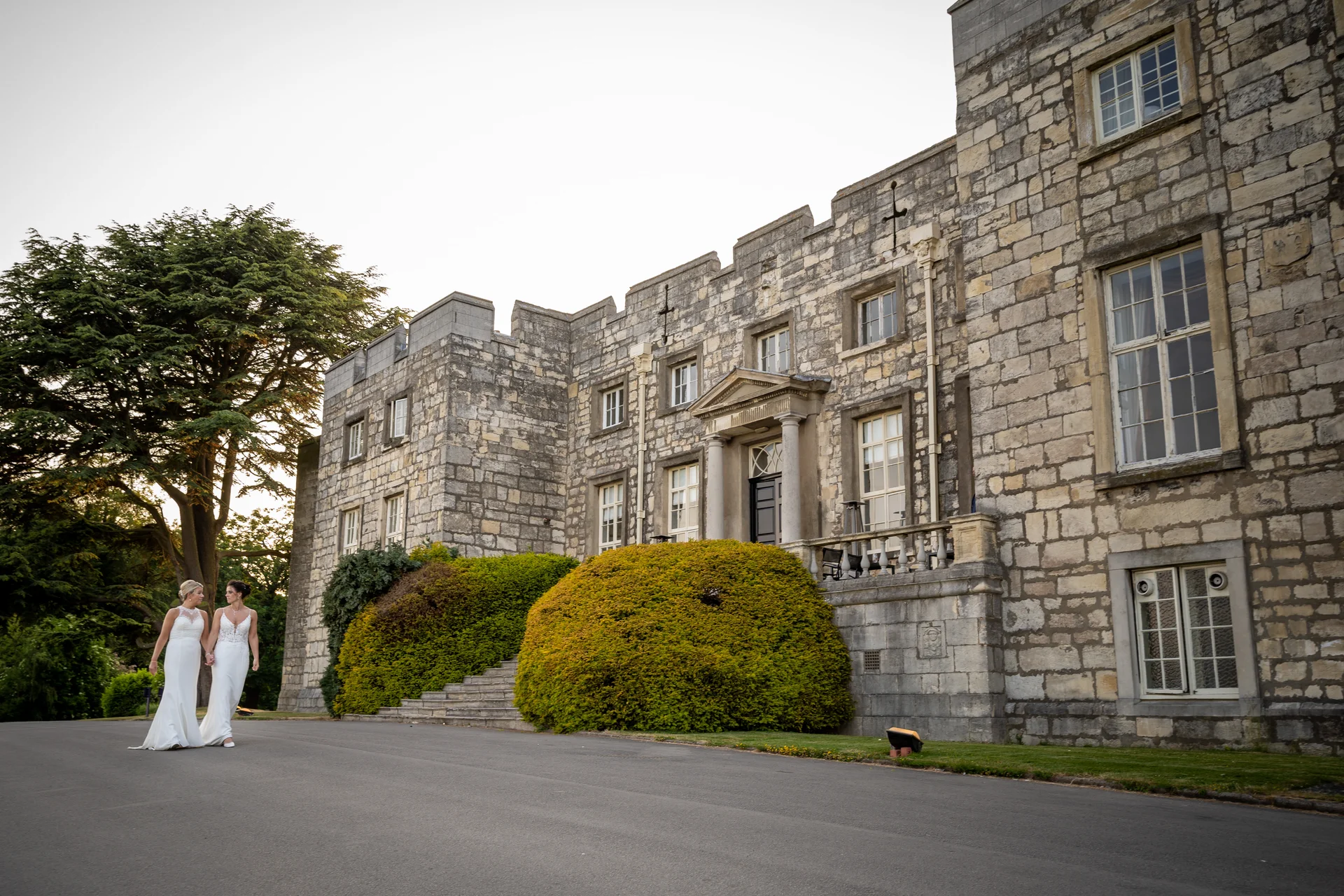 Two brides in white wedding dresses walk hand in hand down the paved driveway in front of a grand historic stone castle.
