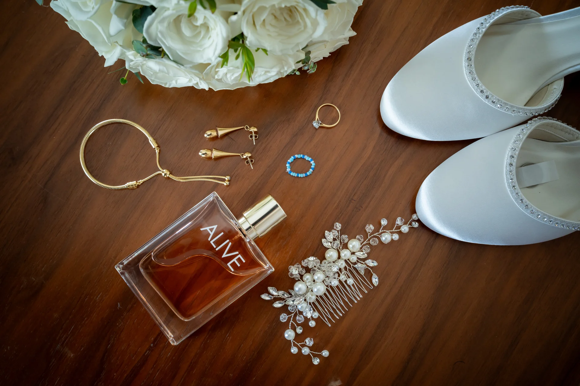 An elegant bridal flat lay showcases white satin crystal-embellished heels, a bottle of perfume, gold jewellery, a hair comb, wedding rings, and a white rose bouquet arranged neatly on a dark wooden surface.