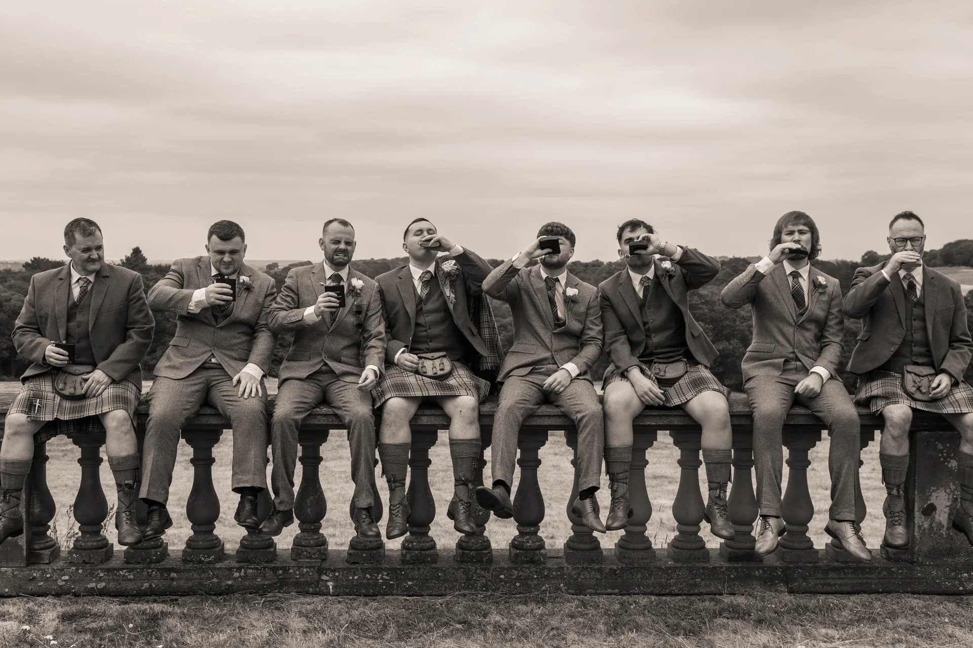 A sepia-toned group portrait of groomsmen in tweed suits and kilts sitting on a stone balustrade, taking sips from pocket flasks against an overcast landscape backdrop.