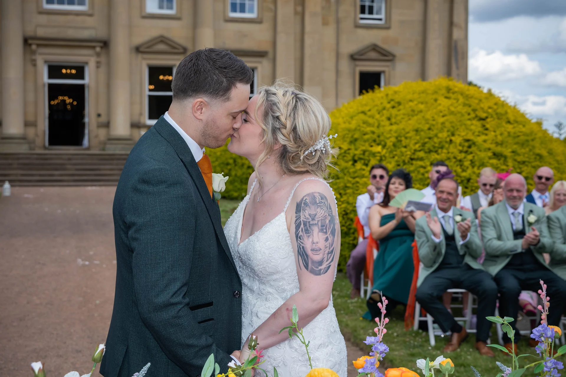A newlywed couple share their first kiss during an outdoor ceremony in front of a grand country house, while seated guests applaud in the background.