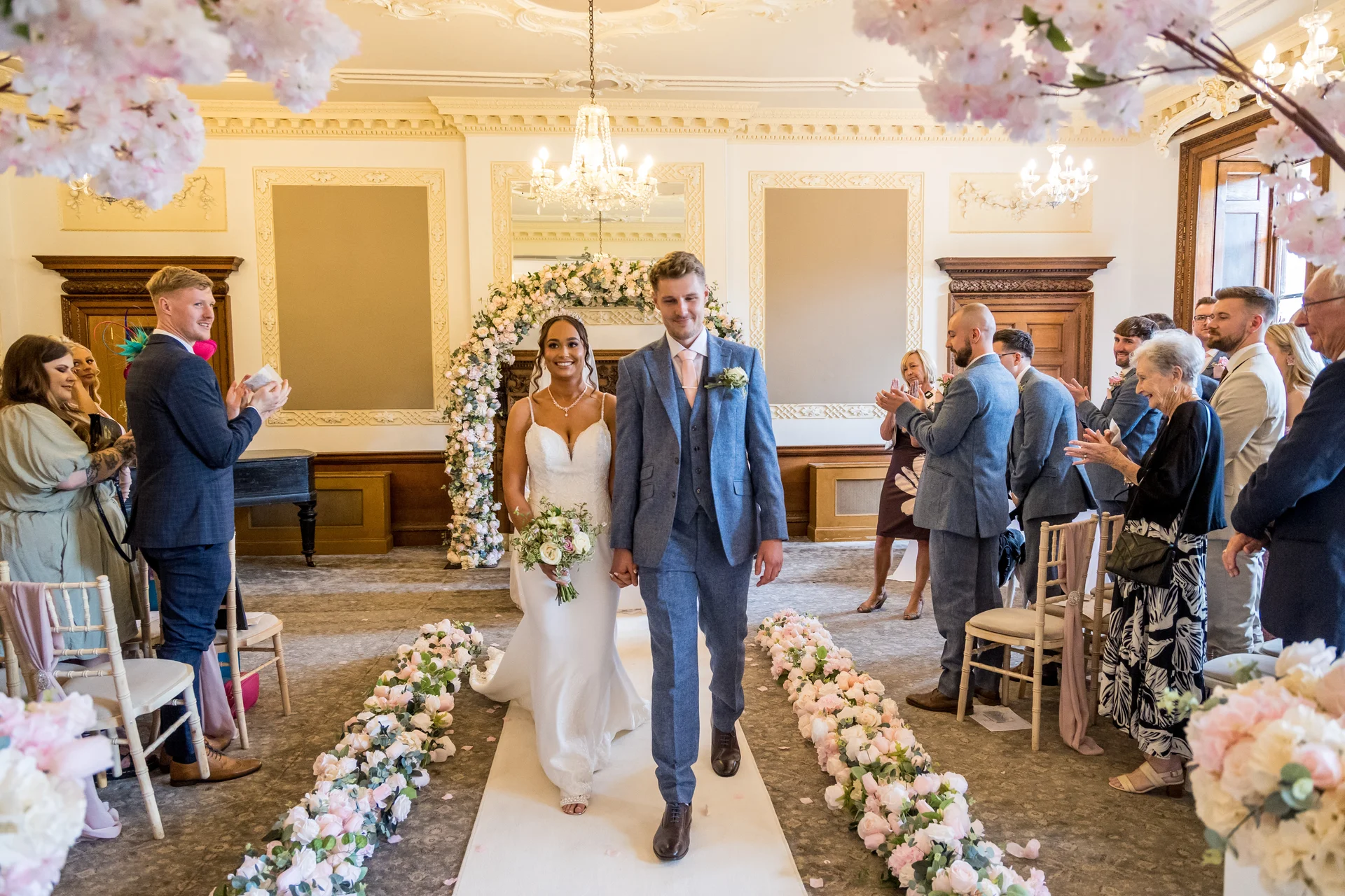 A newlywed bride and groom walk hand-in-hand down the aisle lined with floral arrangements in a grand ballroom as guests cheer and applaud.