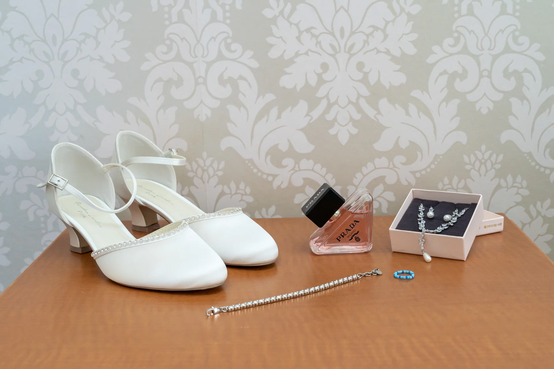 A flat lay arrangement of bridal details on a wooden surface featuring satin block heel shoes, a Prada perfume bottle, jewellery in a presentation box, and a blue beaded ring.