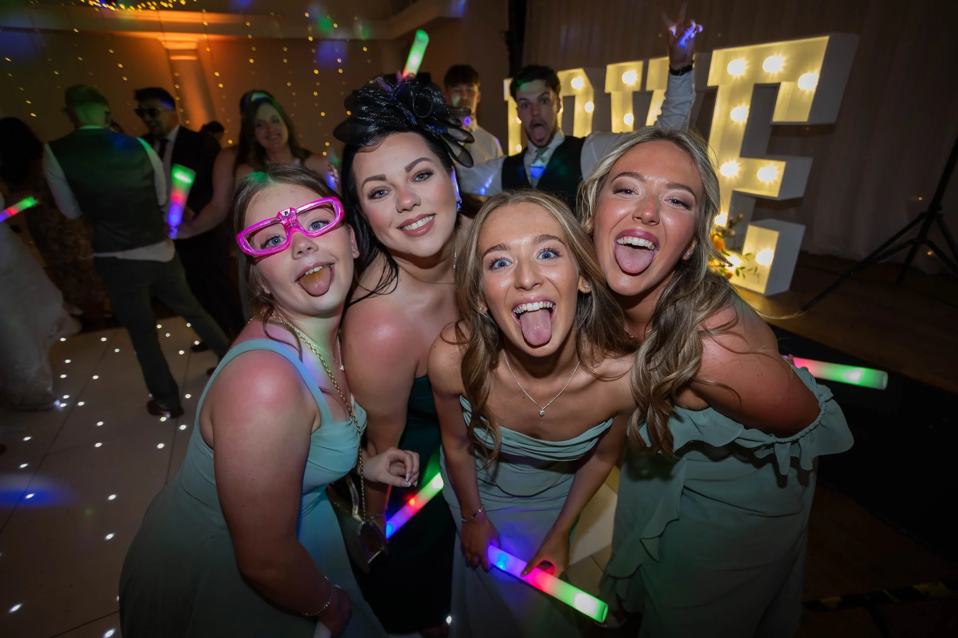 A group of bridesmaids and wedding guests playfully pose on the dance floor holding glow sticks beneath glowing letter lights.