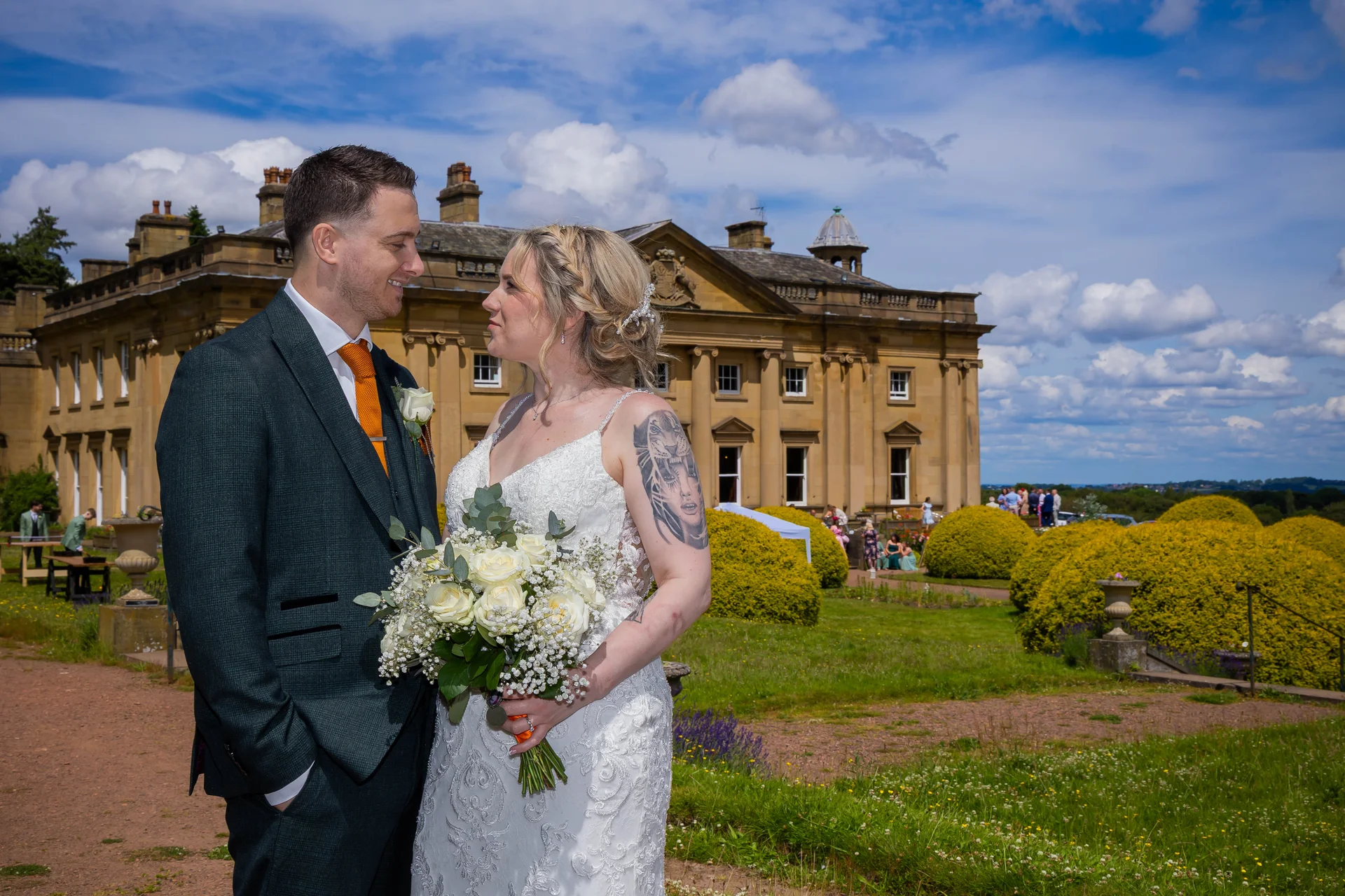 A bride with tattoos and a groom in a tweed suit gaze at each other holding a white rose bouquet outside a grand country house.