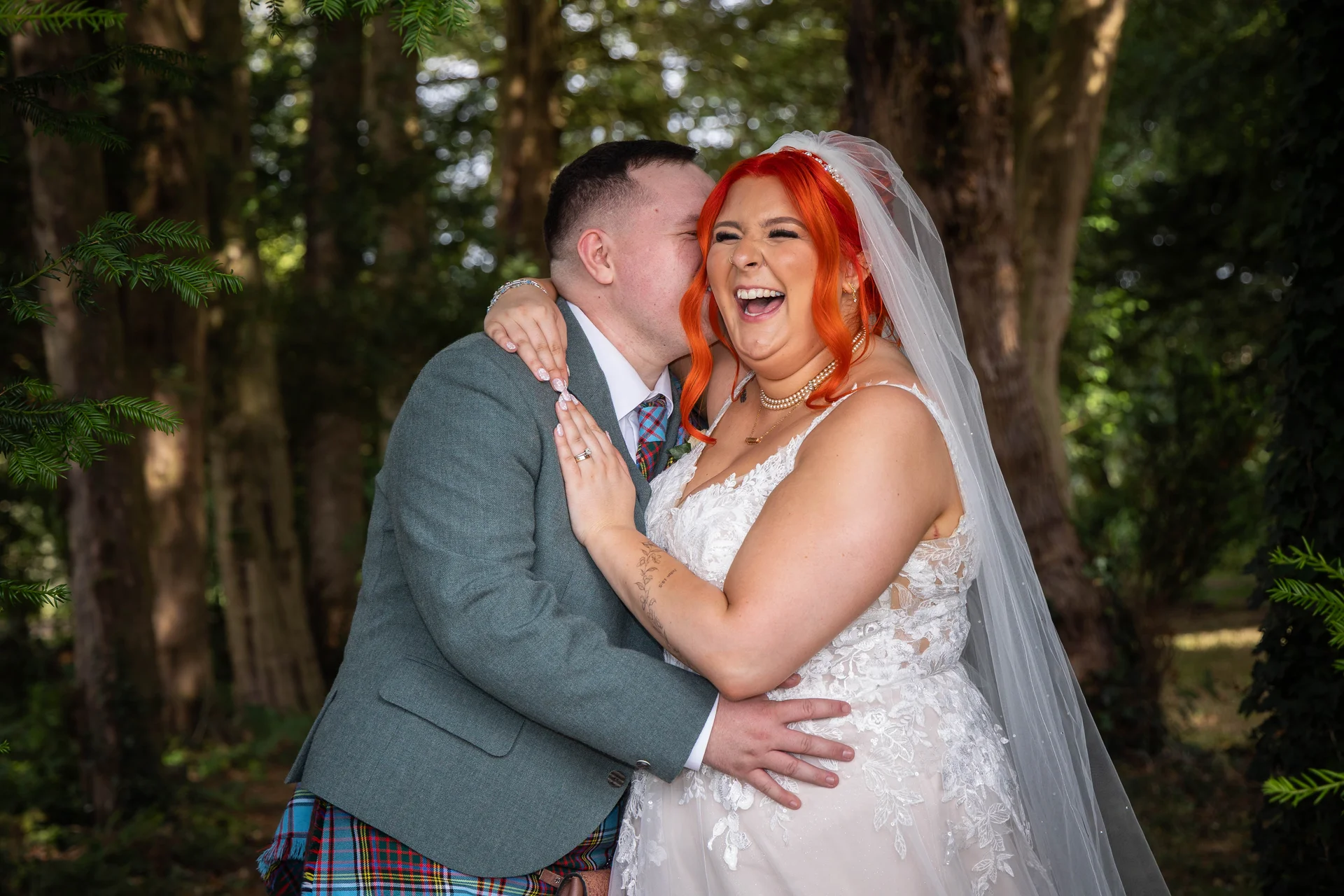A joyful couple embraces in a woodland setting, with the laughing bride wearing a lace wedding dress and bright red hair while her partner wears a kilt and tweed jacket.
