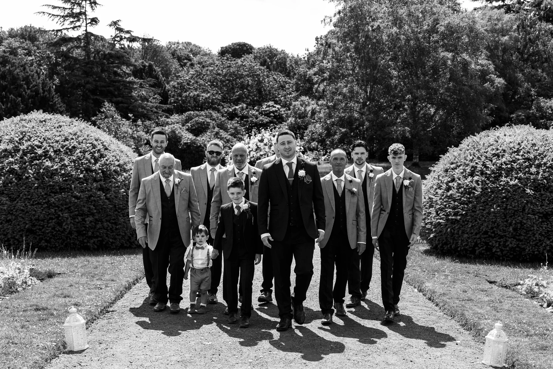 The groom and his groomsmen stride together down a sunlit garden path, flanked by manicured shrubbery in a classic black and white capture.